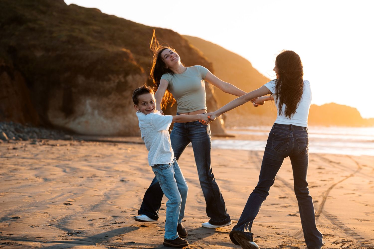 Two women and a young boy playing and dancing together on a sandy beach at sunset.