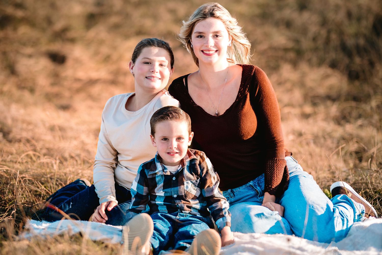 A smiling woman with two children sitting on a blanket in a golden field for a warm family portrait outdoors.