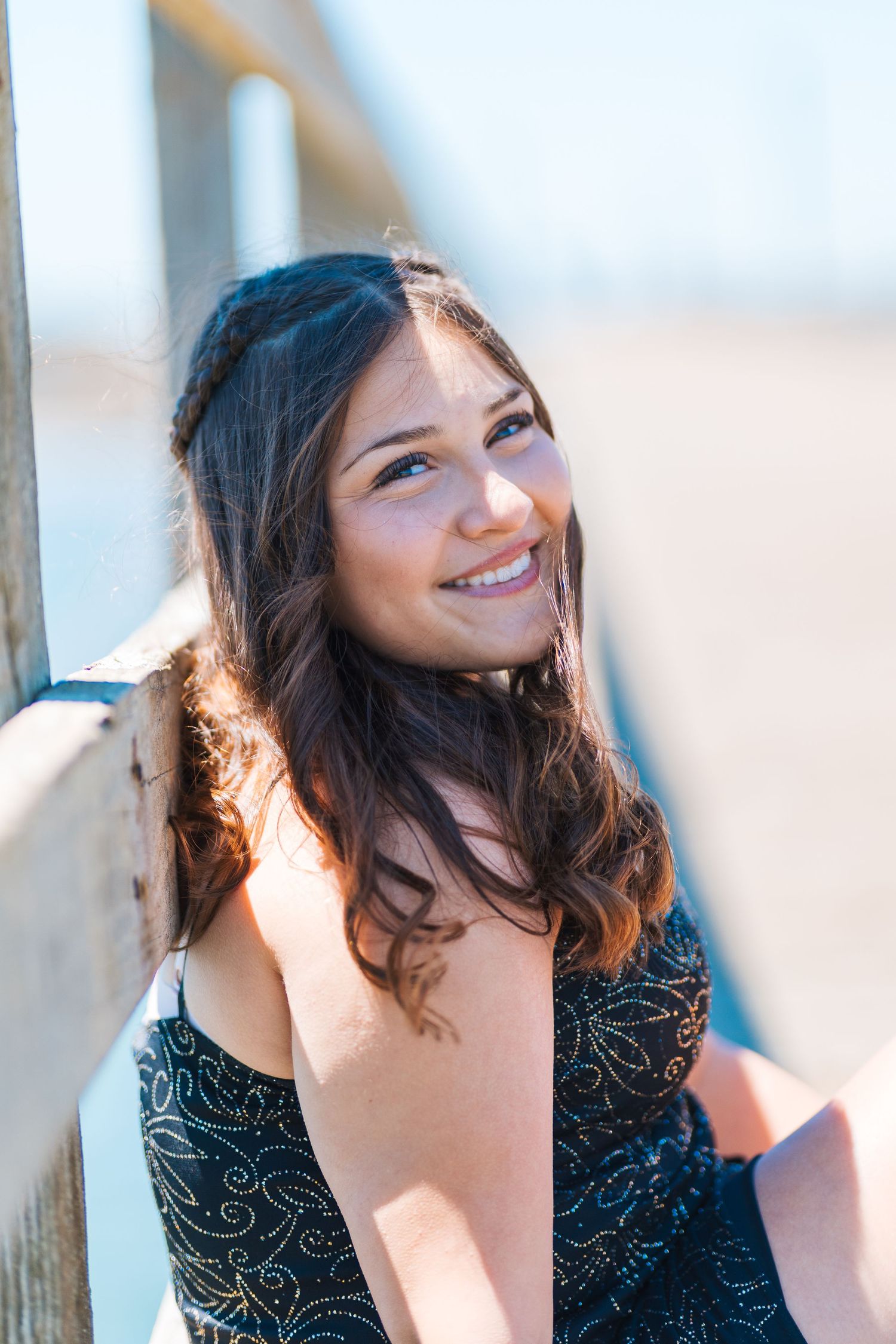 Smiling young woman with long dark hair wearing a black patterned dress, posing outdoors in bright sunlight.