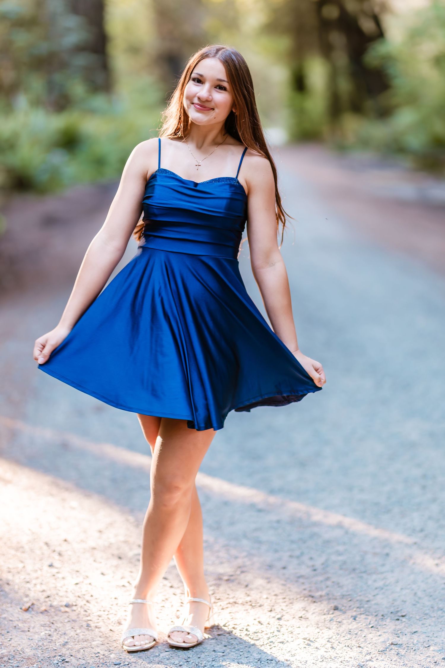 Teen girl twirling in a royal blue satin dress on a sunlit forest road.