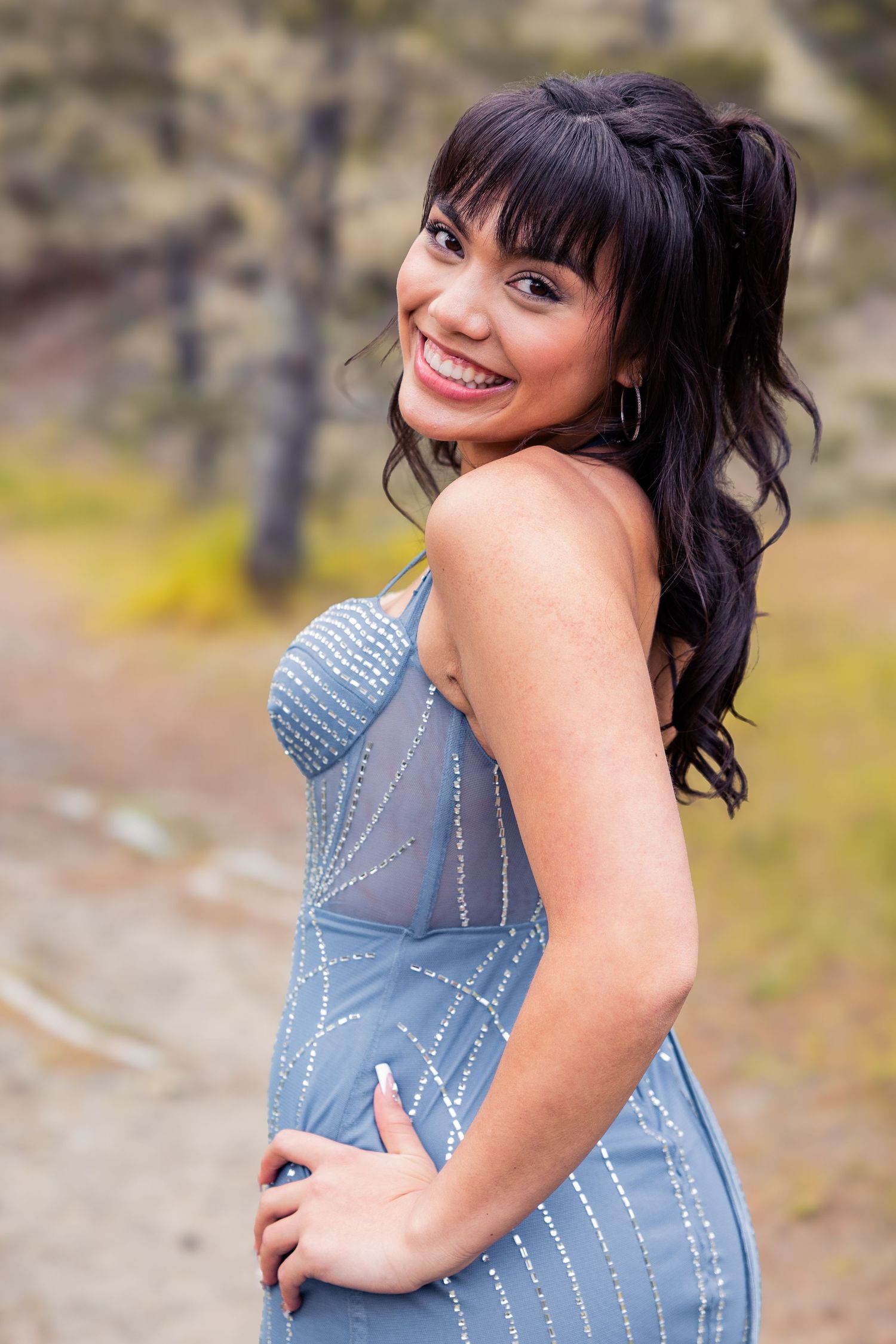 Smiling brunette woman in a blue beaded dress poses outdoors on a dirt path with trees in the background.