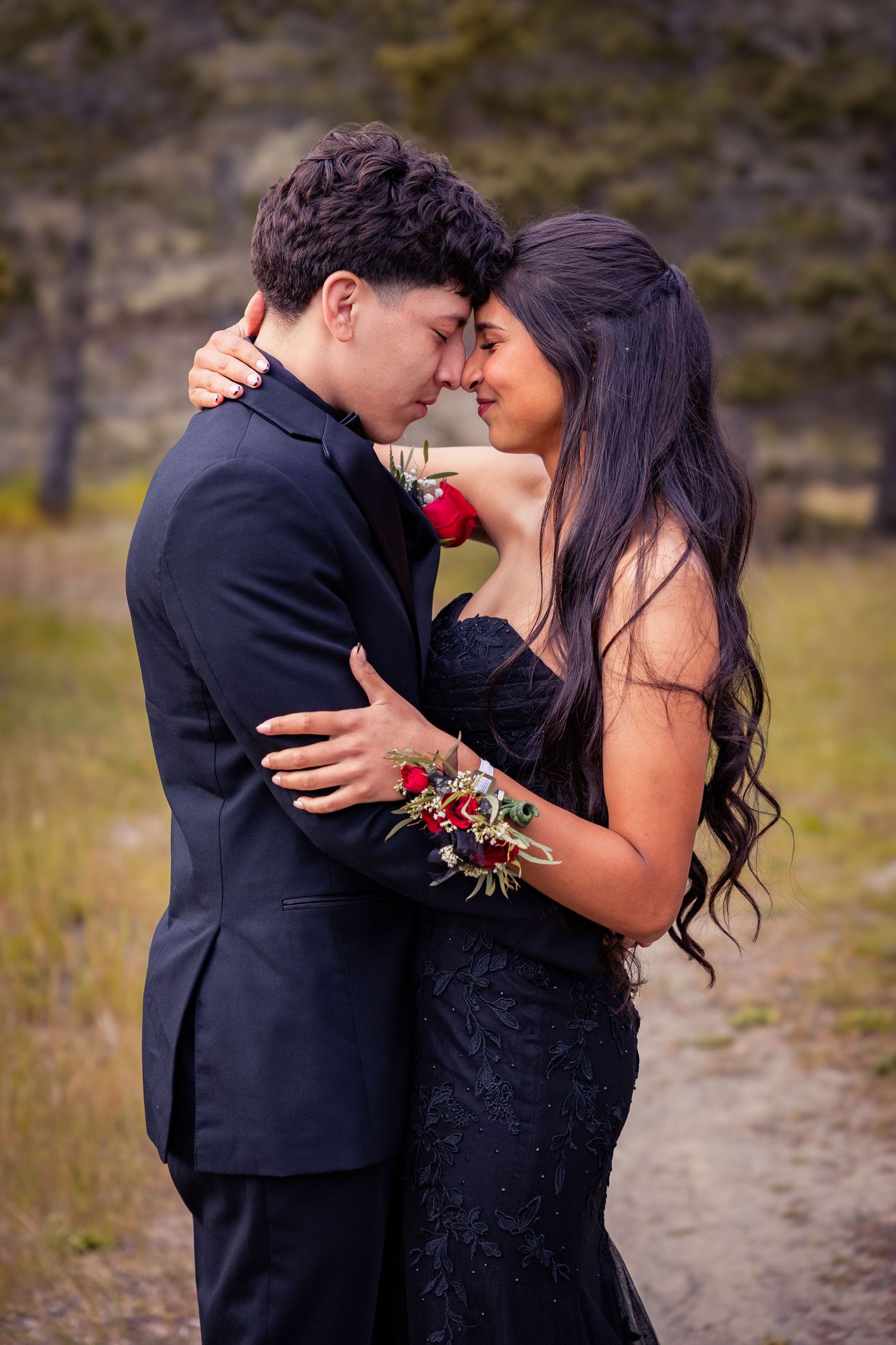 A couple in formal prom attire embrace closely outdoors, wearing matching red floral corsage and boutonniere.