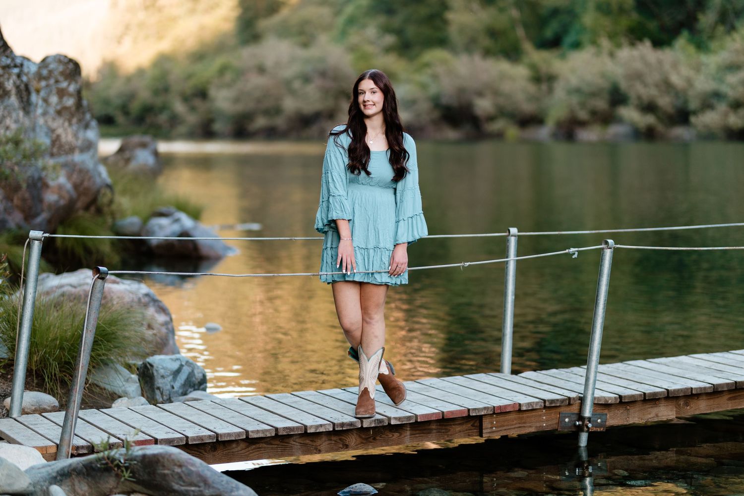 Young woman in light blue dress poses on wooden dock by a serene rocky lake at golden hour.