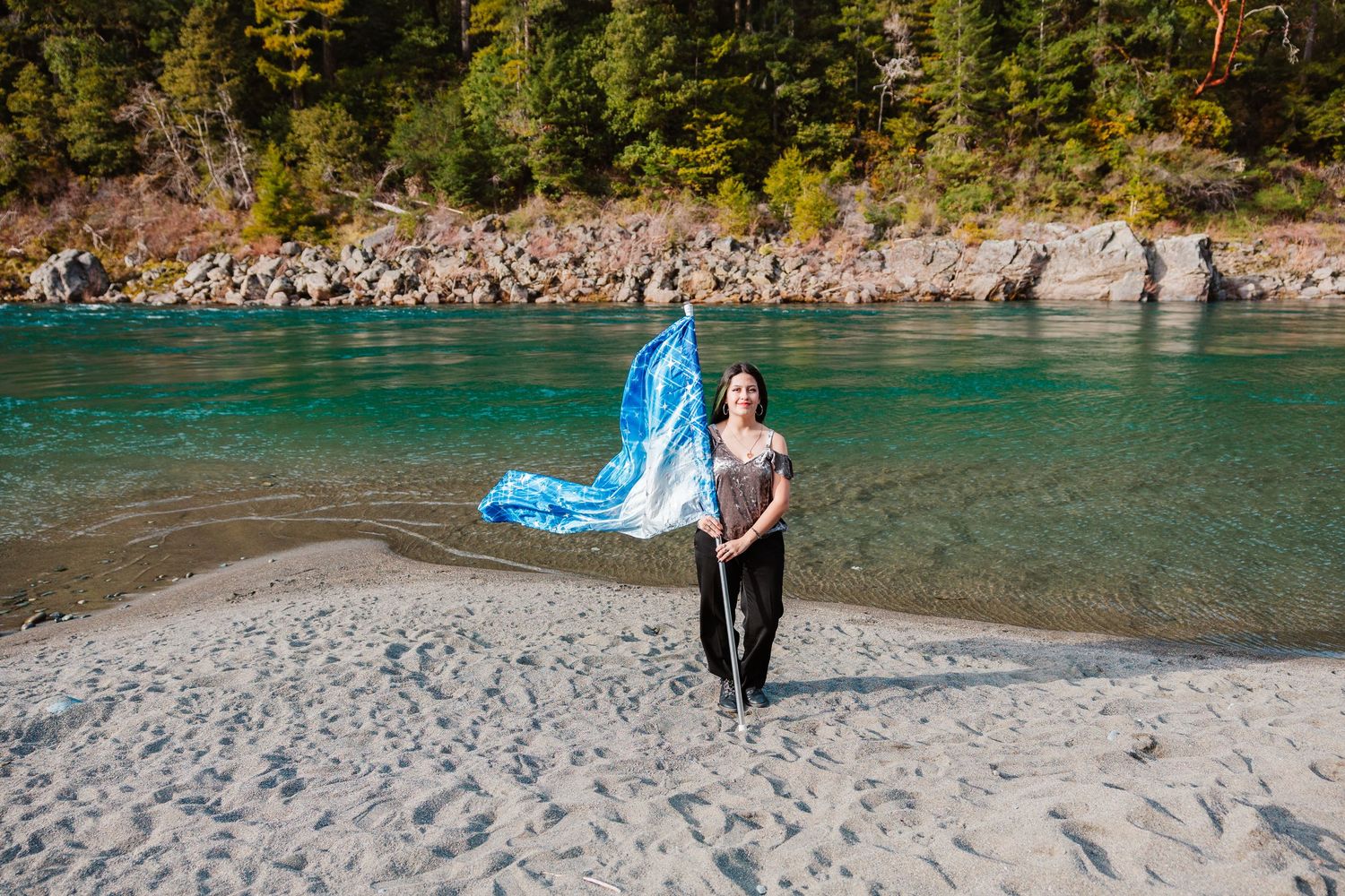 Woman waves a blue and white flag on a sandy riverbank surrounded by turquoise water and trees.