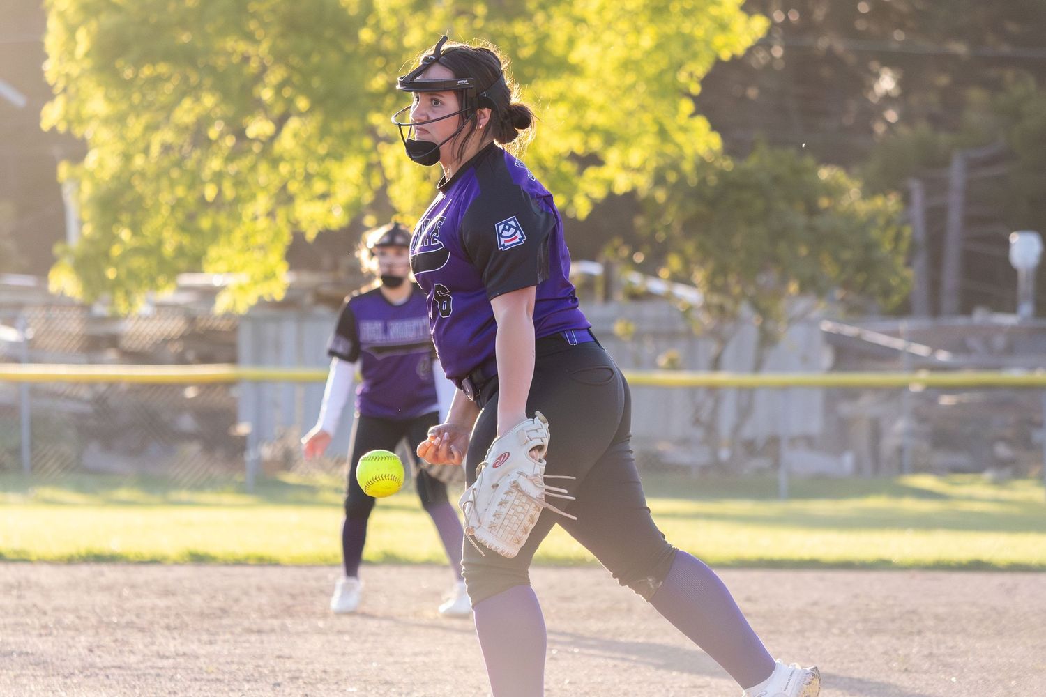 Softball pitcher in purple uniform releases ball during a sunny outdoor game with teammate visible in background.