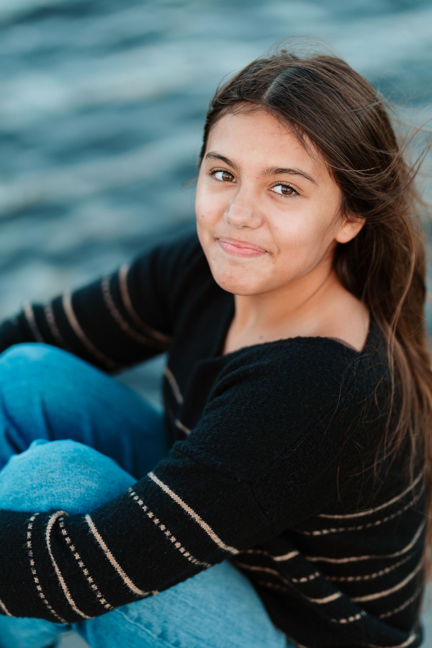 Teenage girl with dark hair smiling while sitting near water wearing a black striped sweater and jeans.