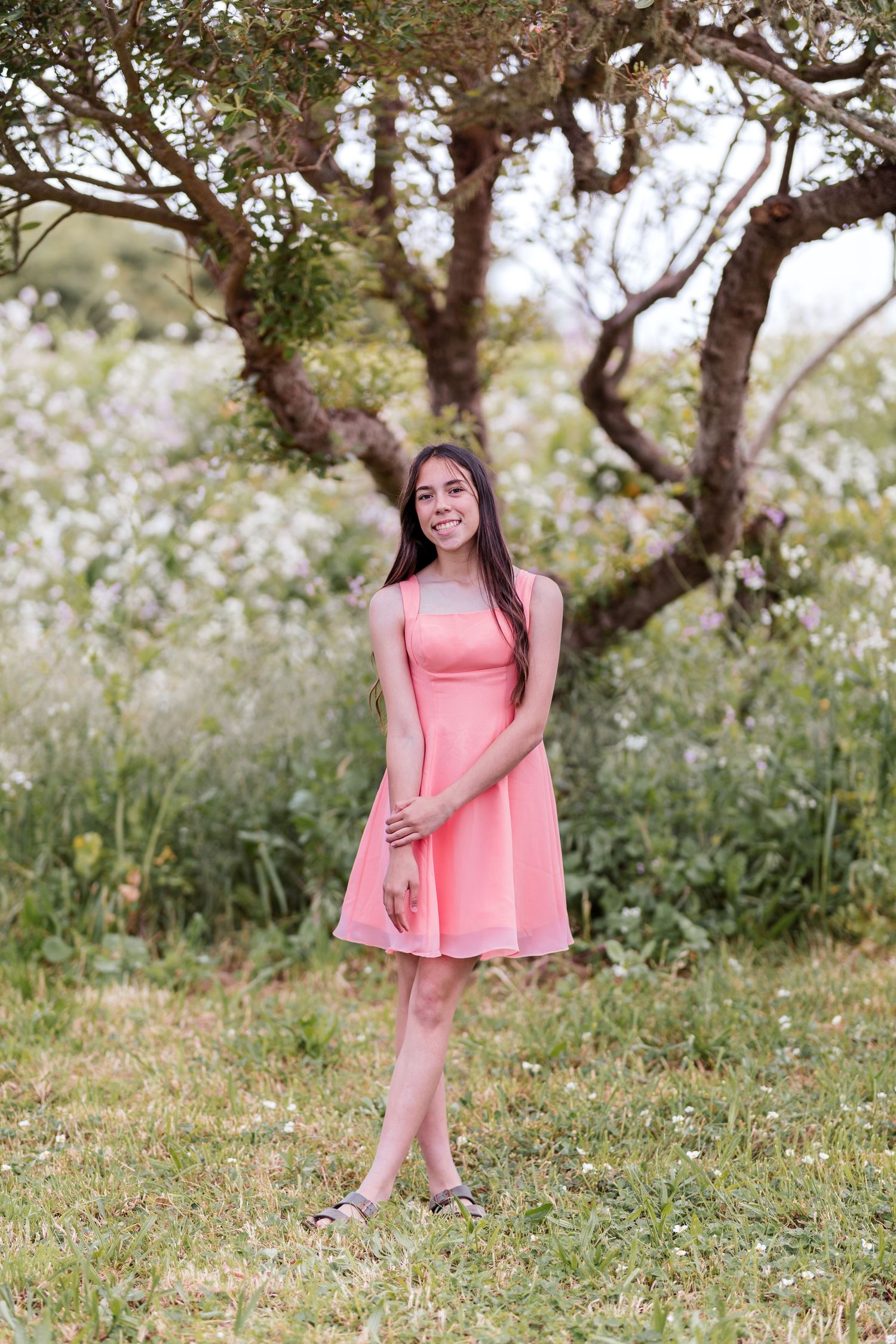 Young woman in pink dress smiling outdoors under a tree with white wildflowers in background.