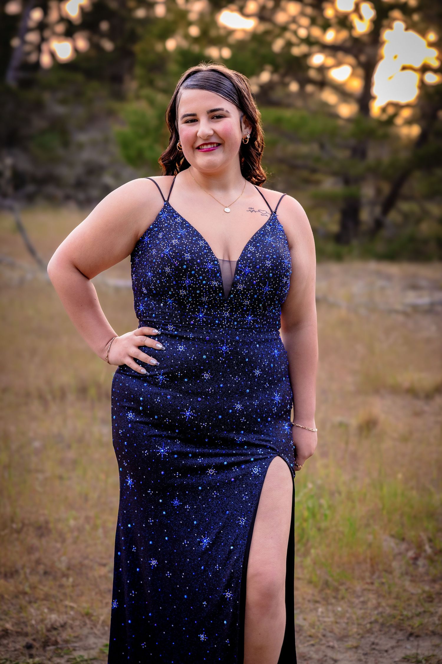 Young woman in a navy blue star-print sequin prom dress with spaghetti straps poses outdoors at sunset.