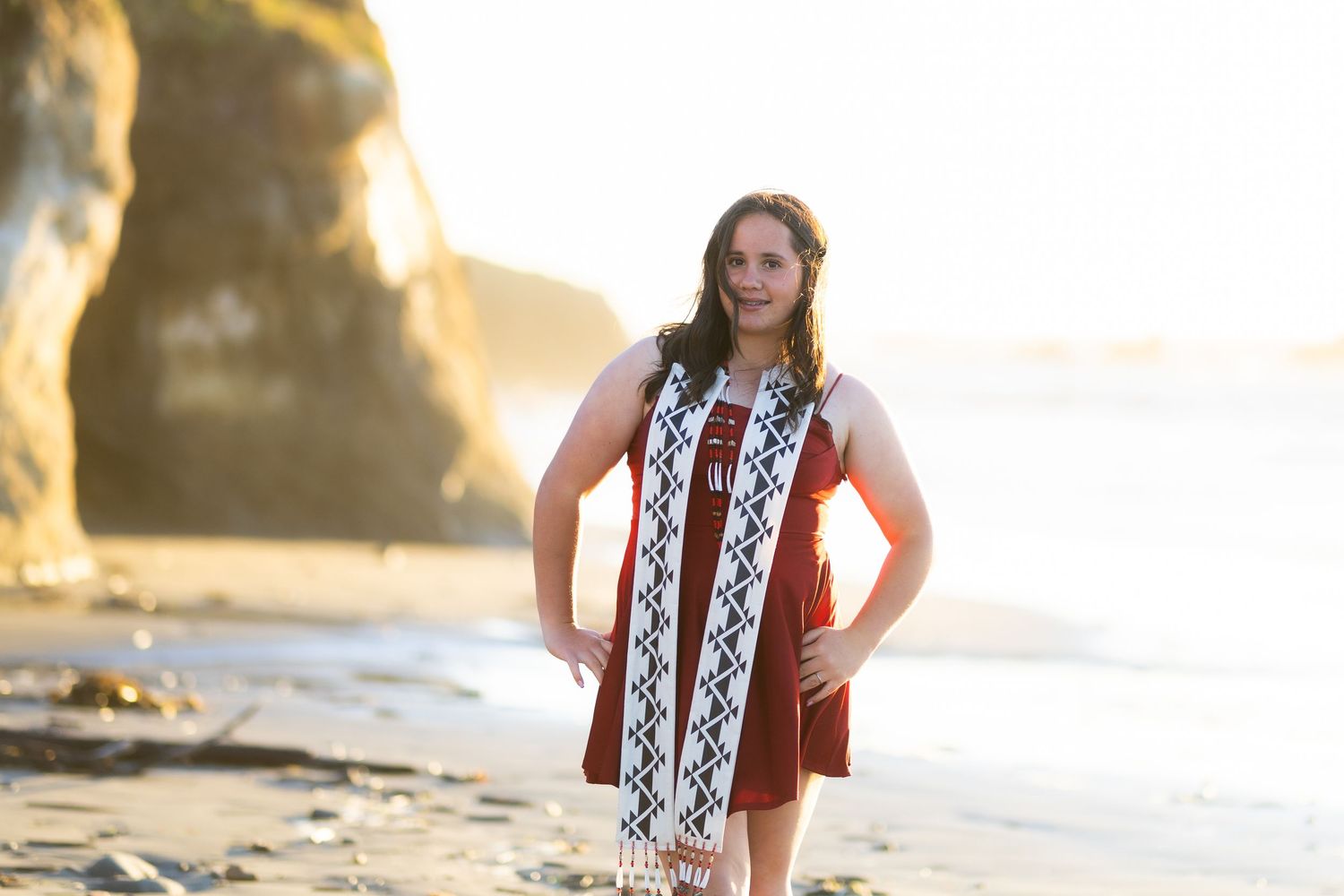 Young woman in maroon dress with patterned sash posing on beach at golden hour.