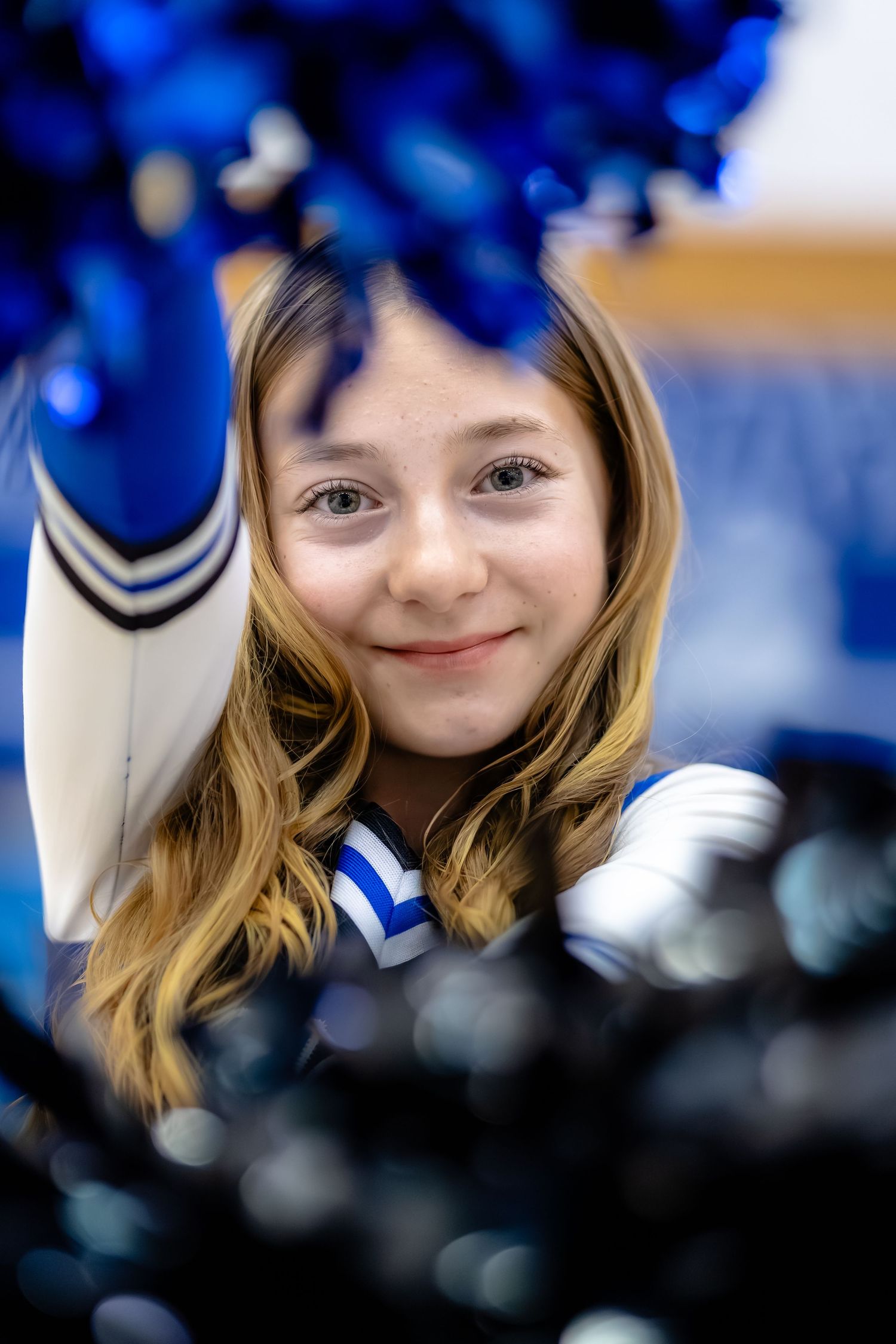 Smiling young cheerleader in blue and white uniform holding blue pom-poms up close to camera.