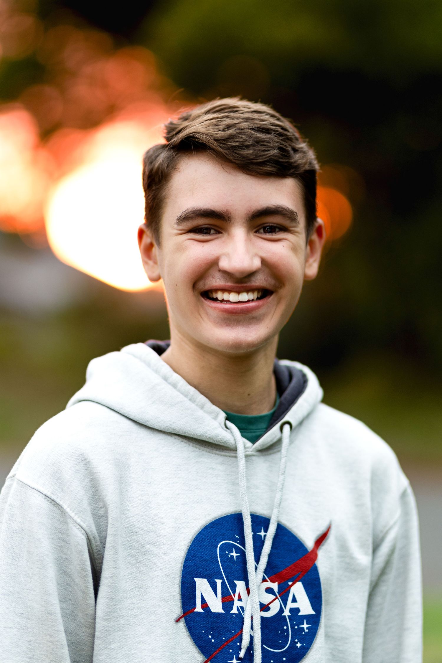 Smiling teenage boy wearing gray NASA hoodie sweatshirt outdoors at sunset.