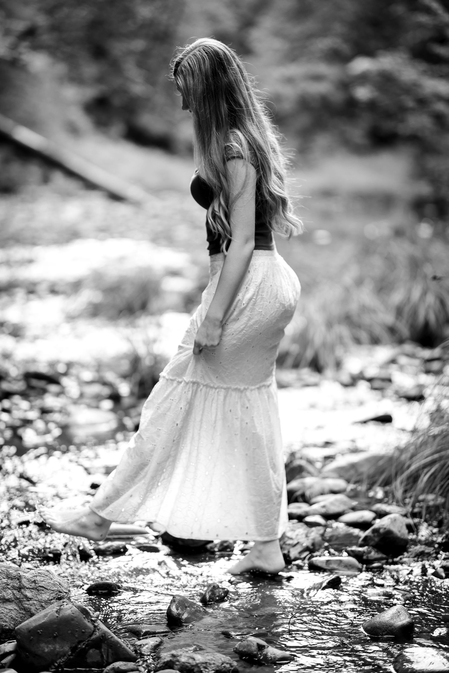 Black and white photo of a woman in a white maxi skirt wading through a rocky forest creek.