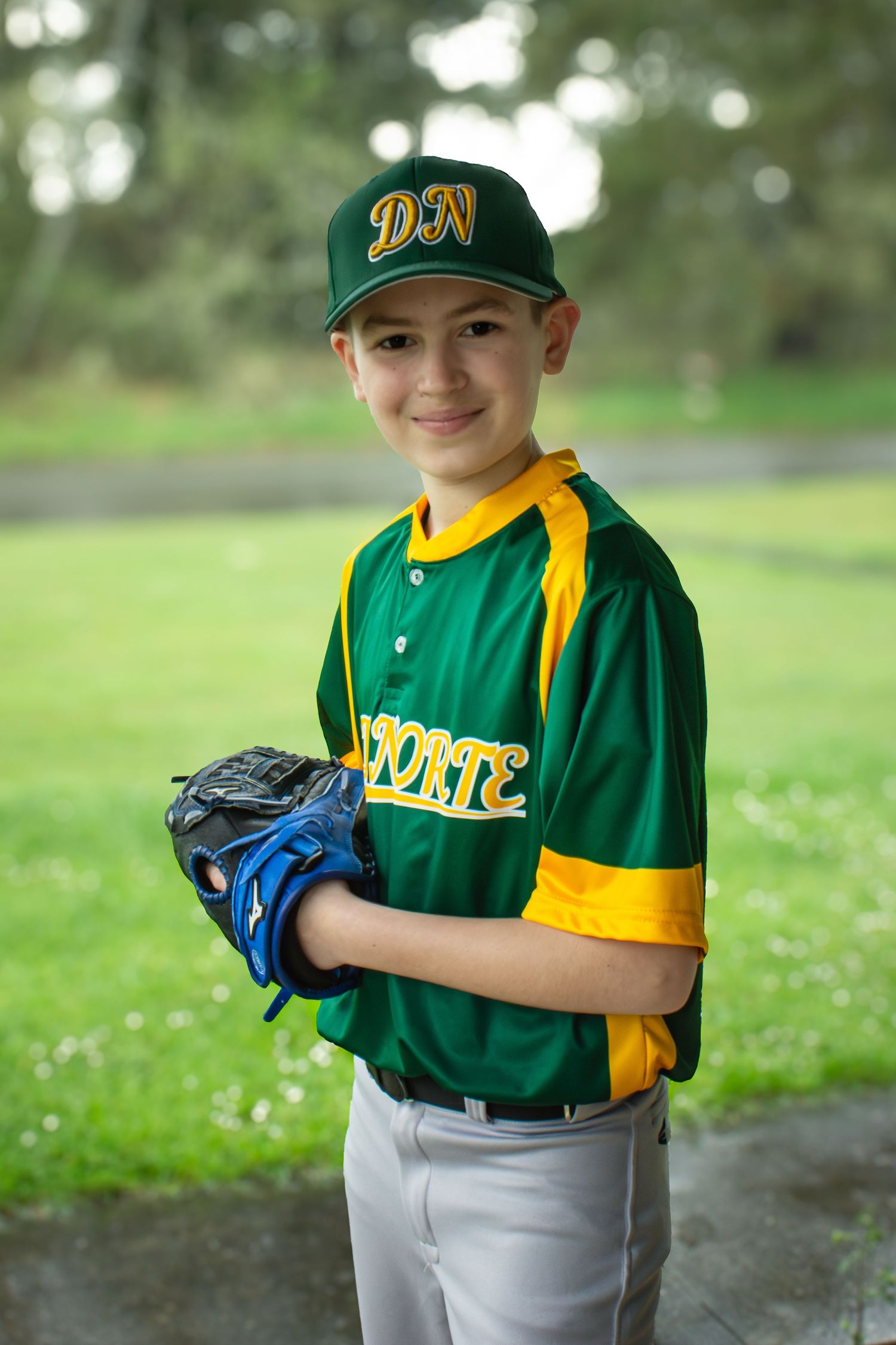 Smiling boy in green and gold DN Norte baseball uniform holding blue glove outdoors.
