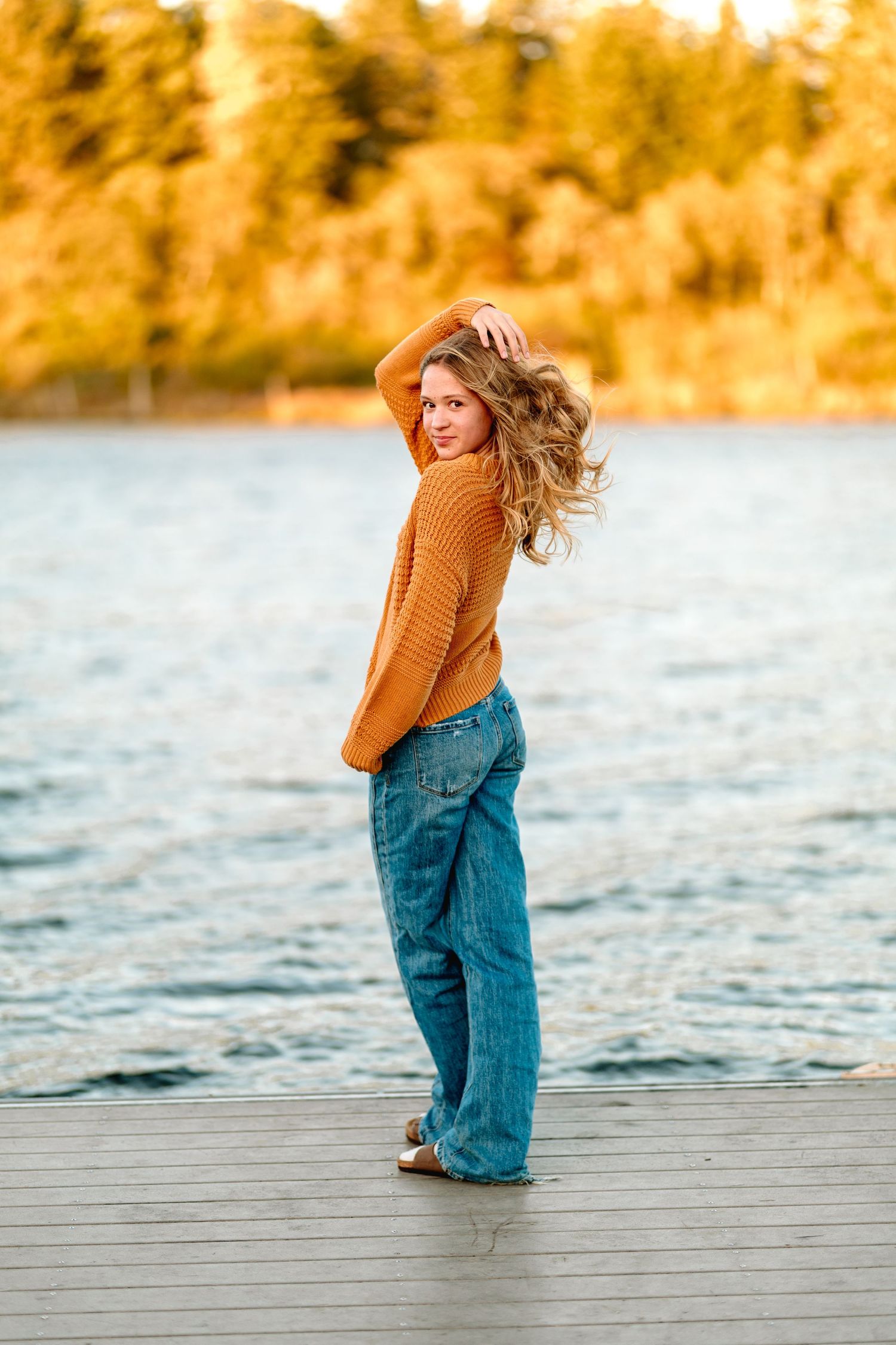 Young blonde woman in orange sweater and wide-leg jeans posing on a lakeside dock at golden hour.