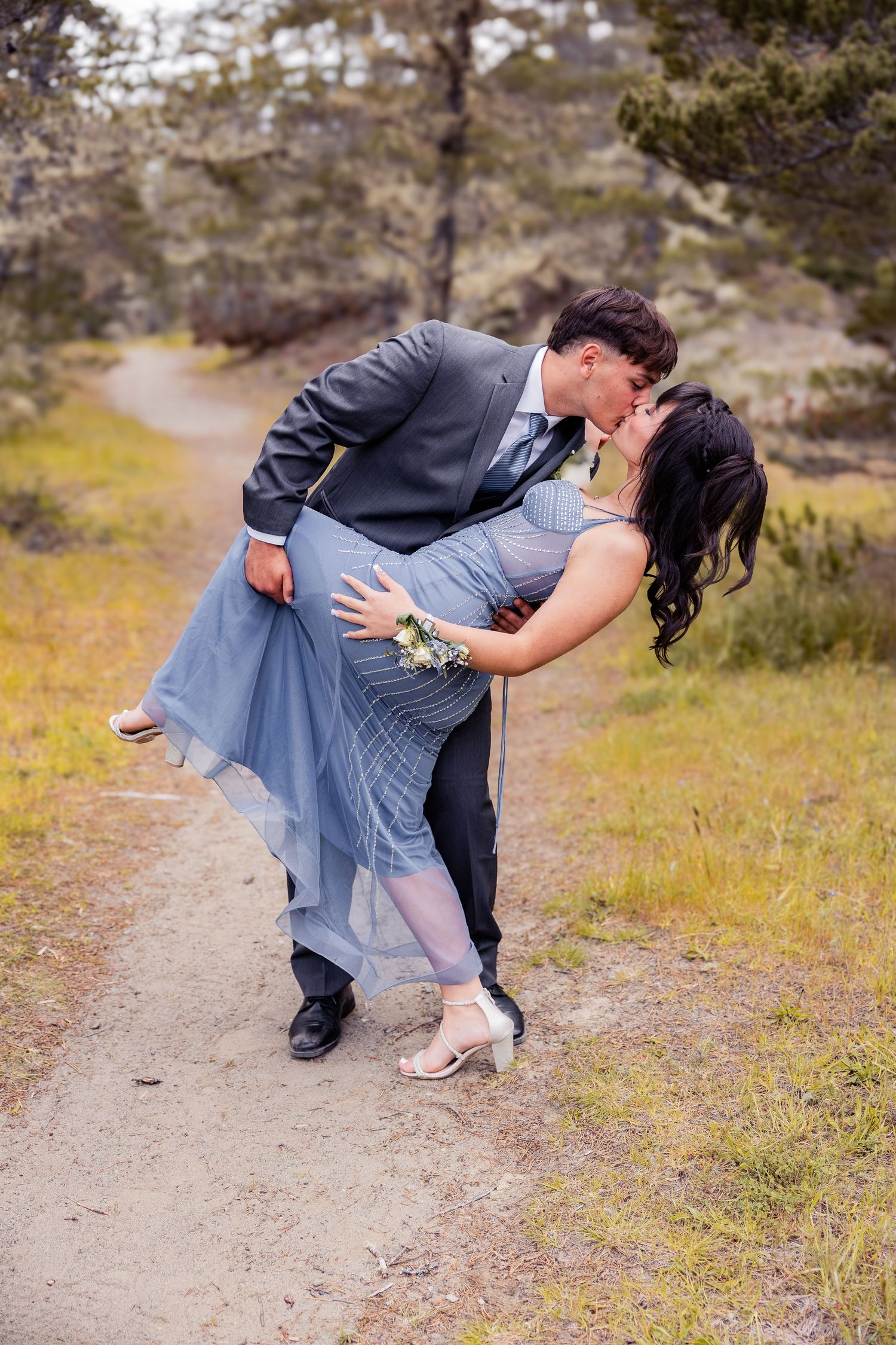Couple sharing a romantic dip kiss on a nature trail, wearing prom attire with a blue gown and gray suit.