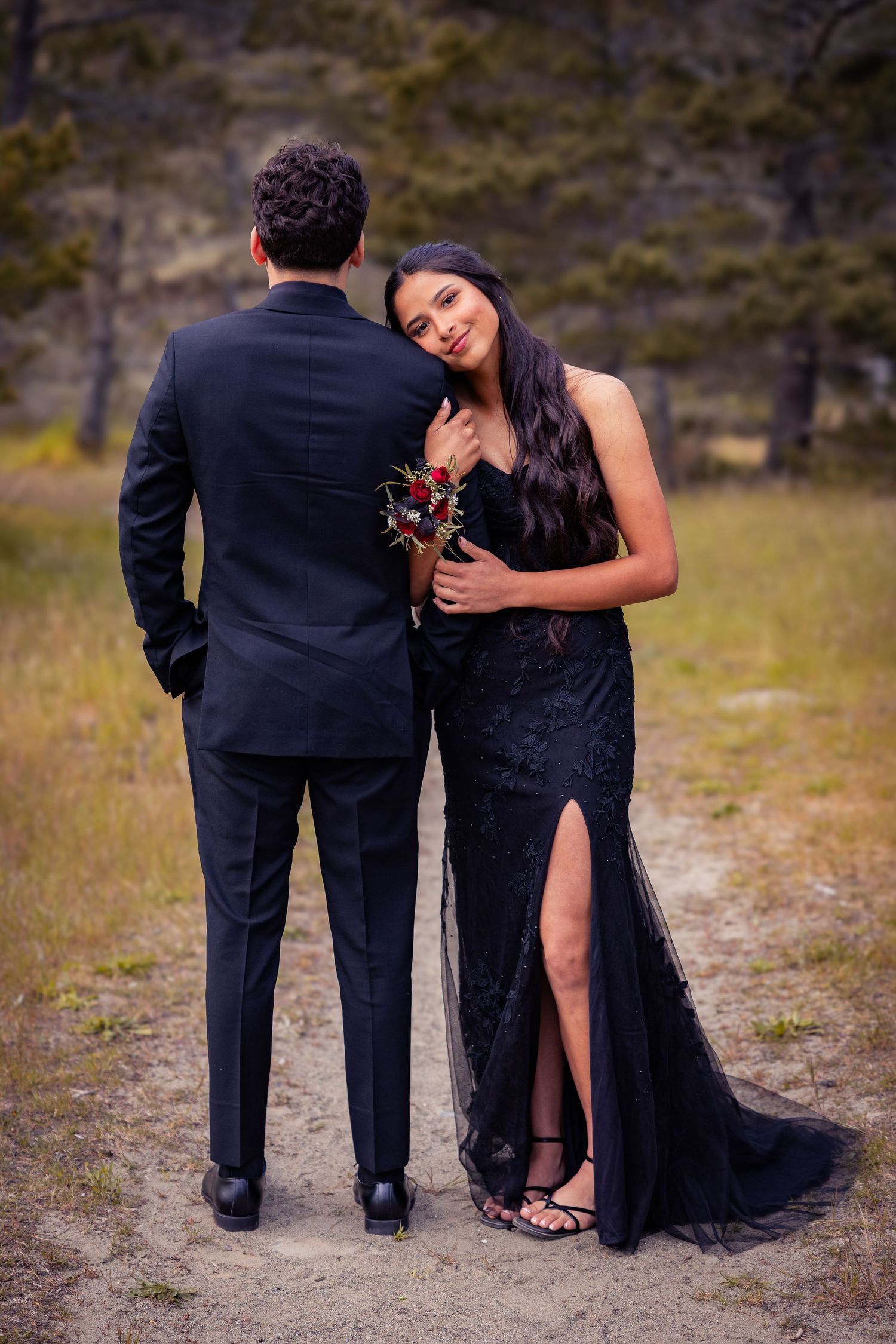Couple in matching black formal wear poses outdoors, woman in lace gown holds bouquet while leaning on mans shoulder.