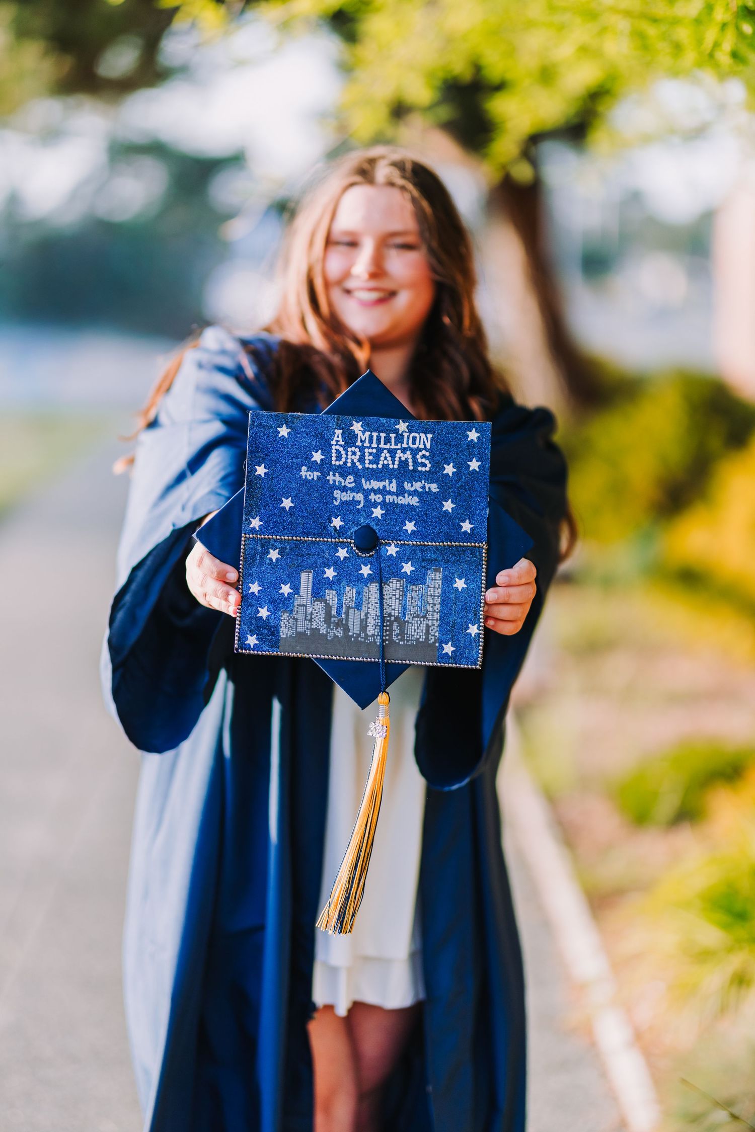 Smiling graduate in blue gown holds decorated cap reading A Million Dreams with city skyline and stars design.