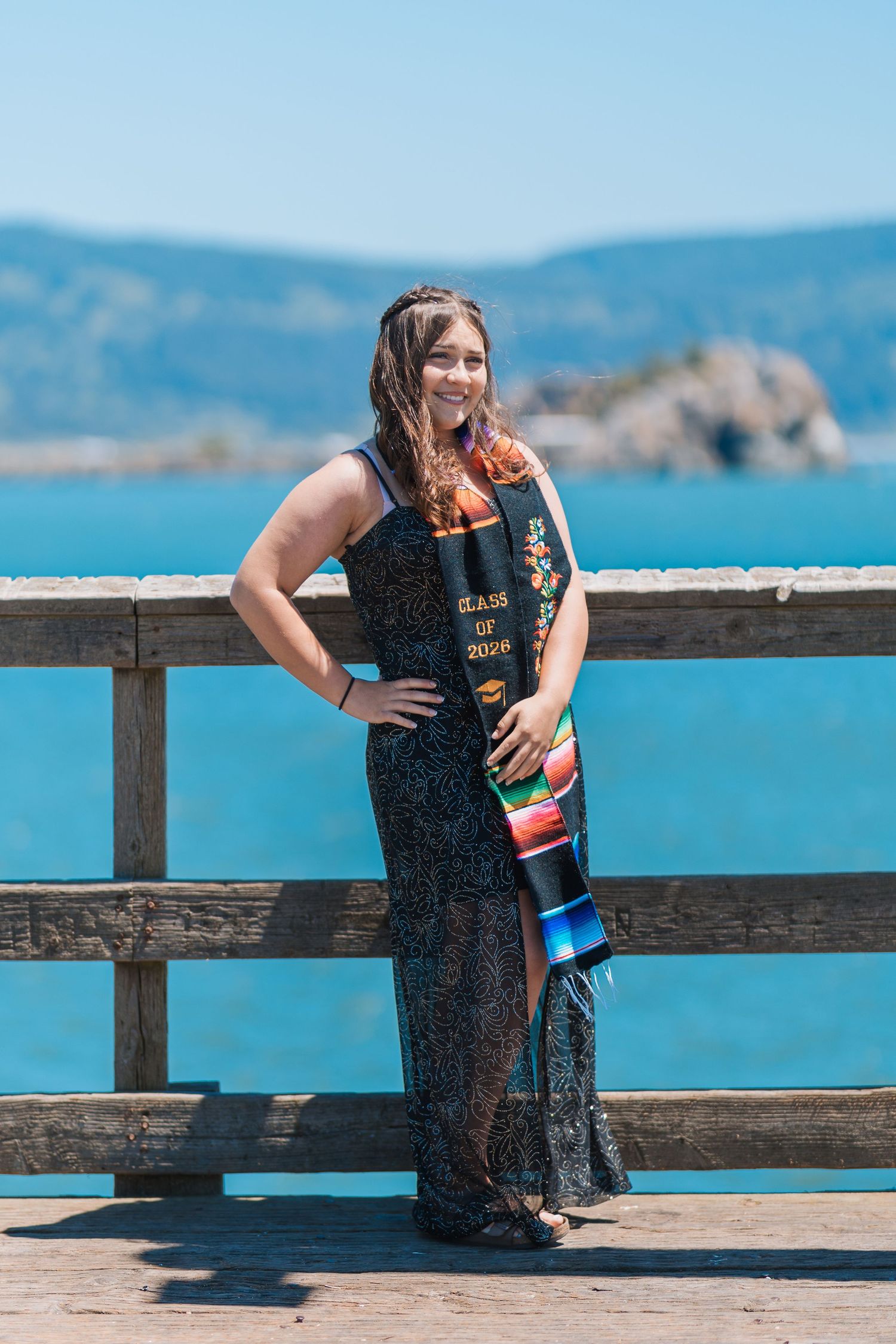 Smiling graduate in black gown holding diploma on a sunny pier with blue ocean and island in background.