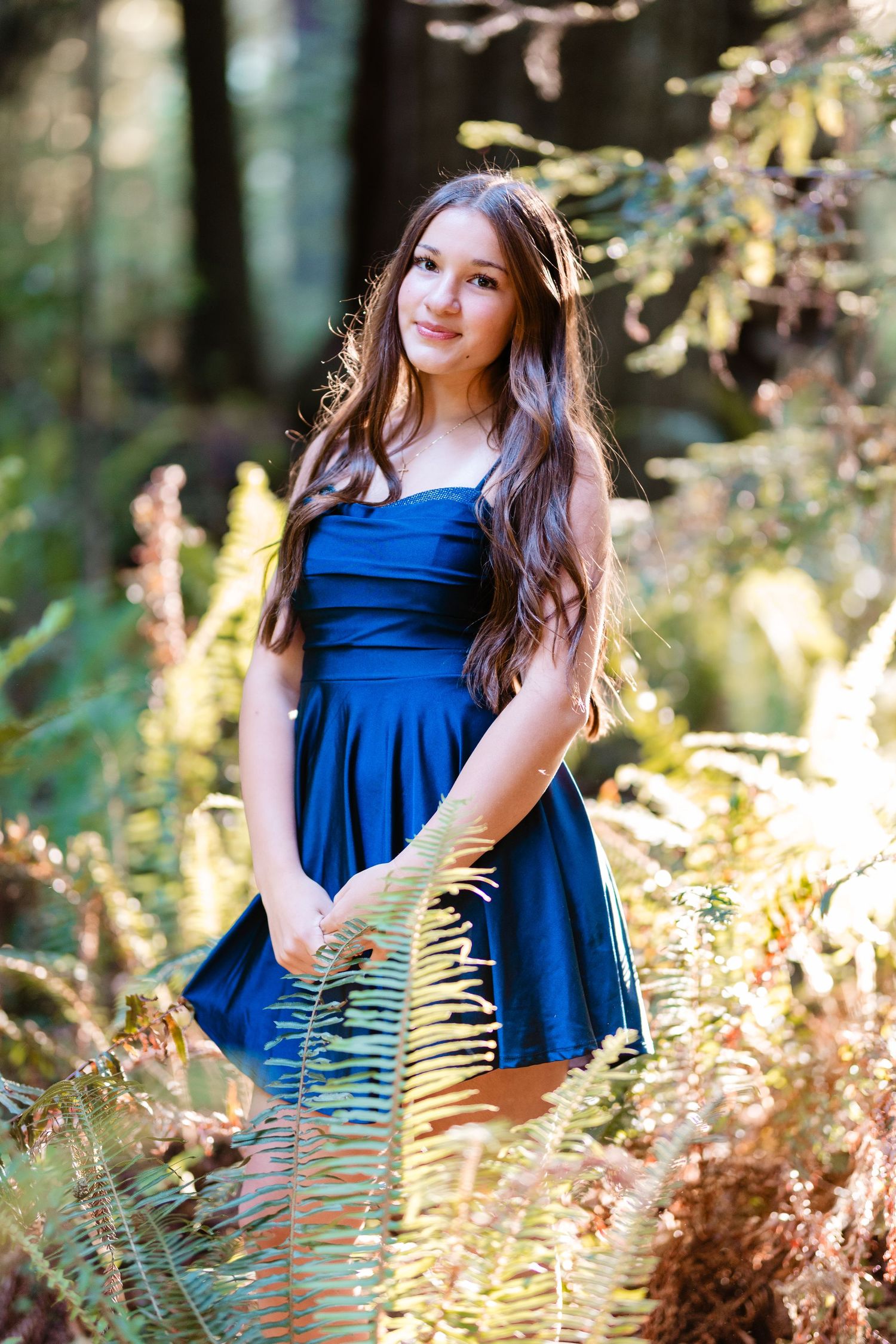Young woman in blue dress standing among ferns in sunlit forest, long dark hair, smiling softly.