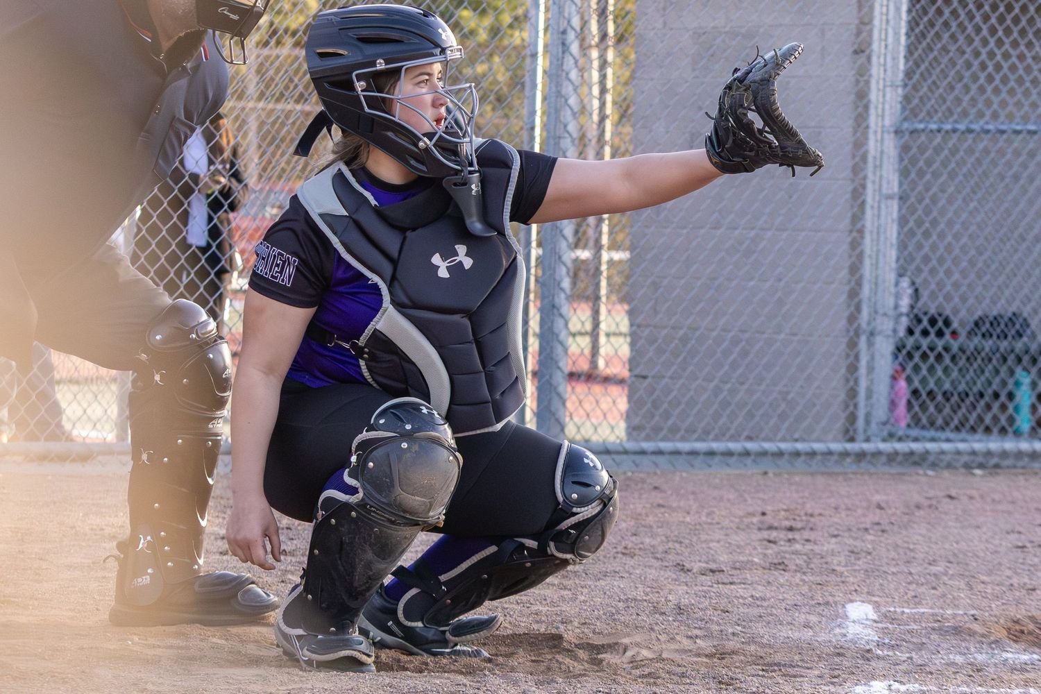 Female softball catcher in full gear crouches behind home plate signaling during a competitive game.