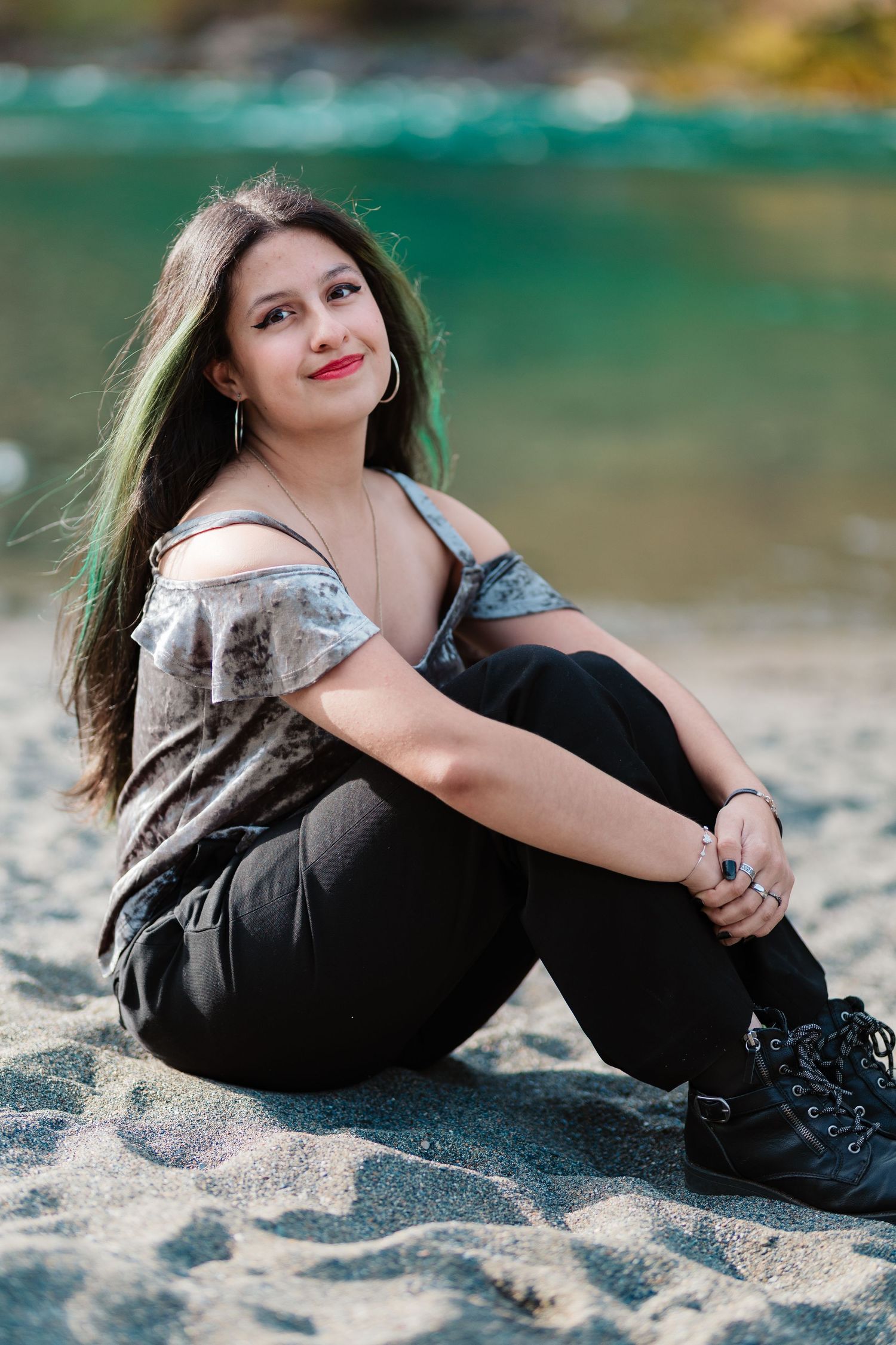 Young woman with long dark hair sits on sandy beach smiling, wearing black pants and gray top.