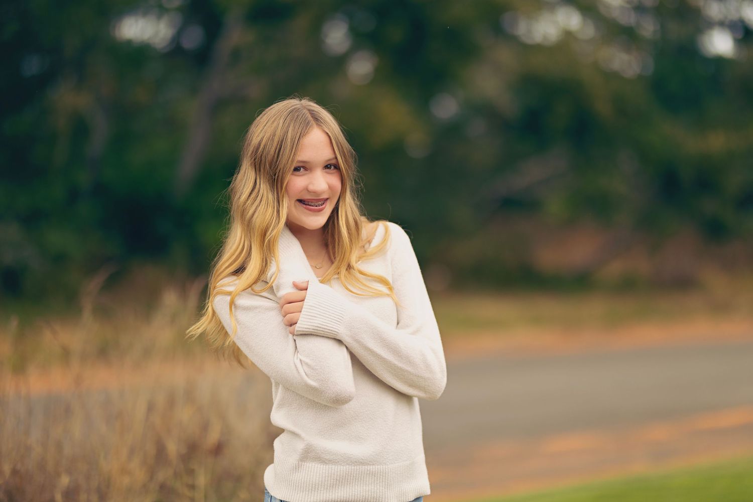 Blonde girl in cream sweater smiling outdoors near road with trees behind.
