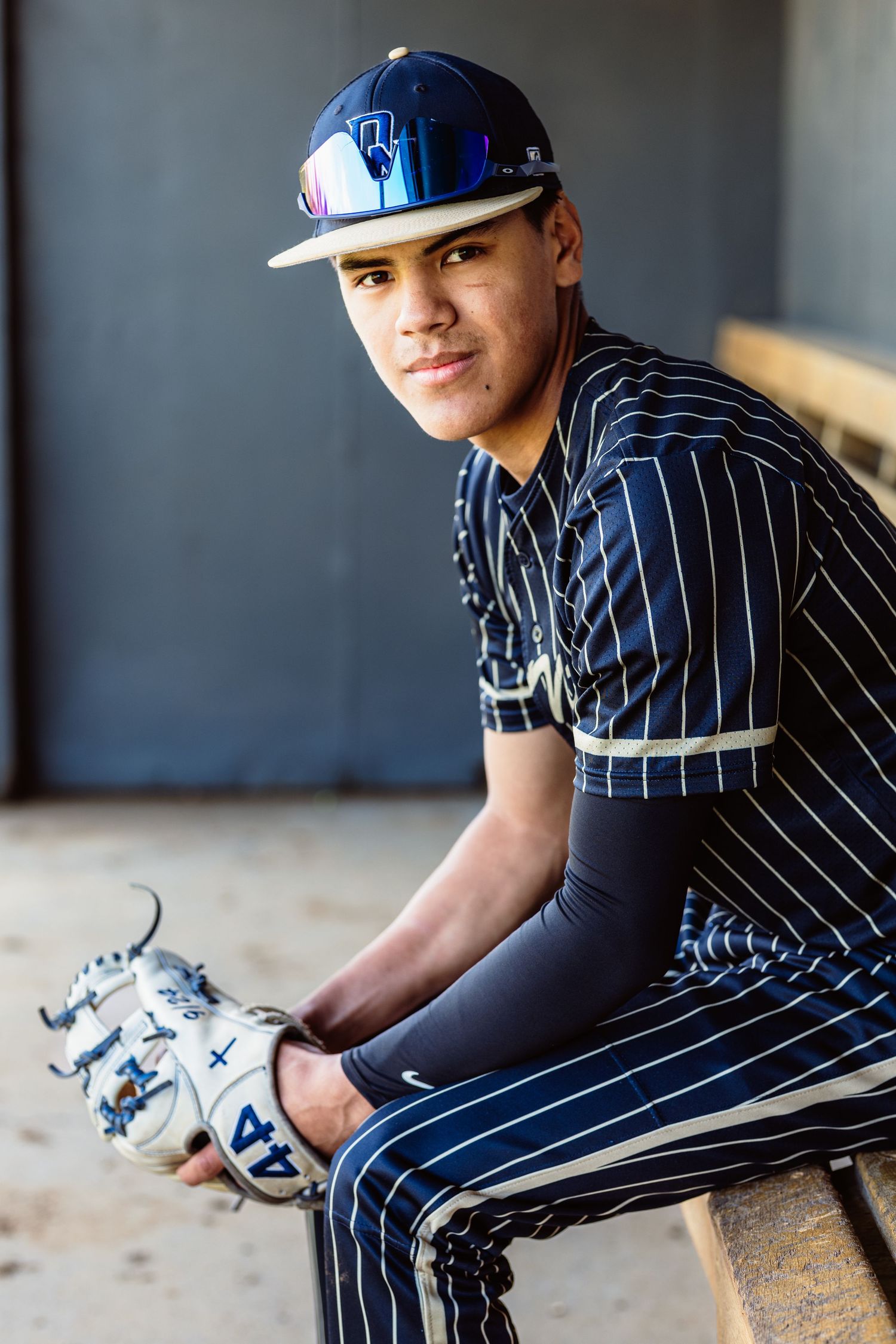 Young baseball player in dark pinstripe uniform sitting in dugout holding glove, wearing blue cap.