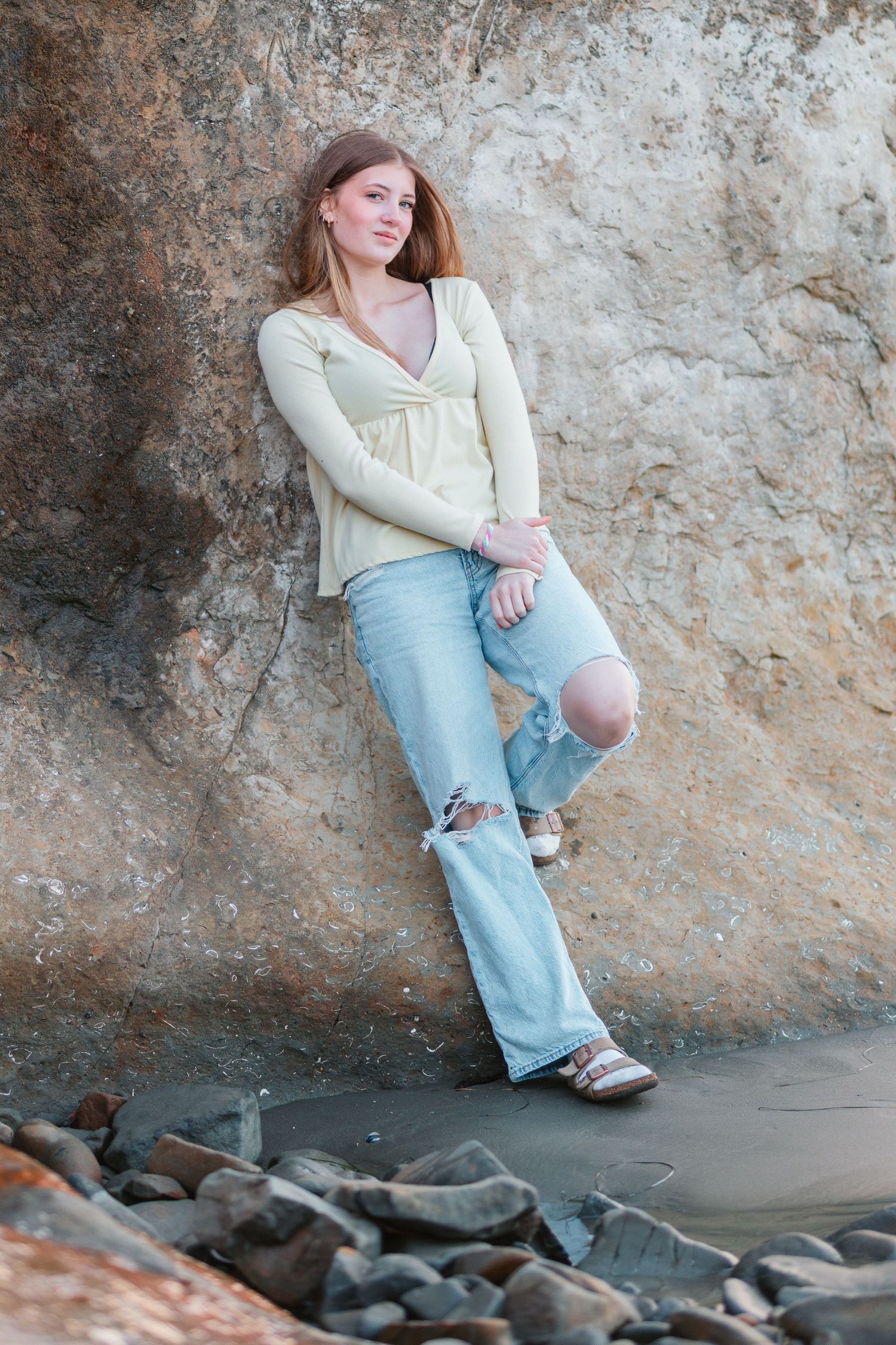 Young woman in cream top and ripped jeans leaning against a large rock formation.