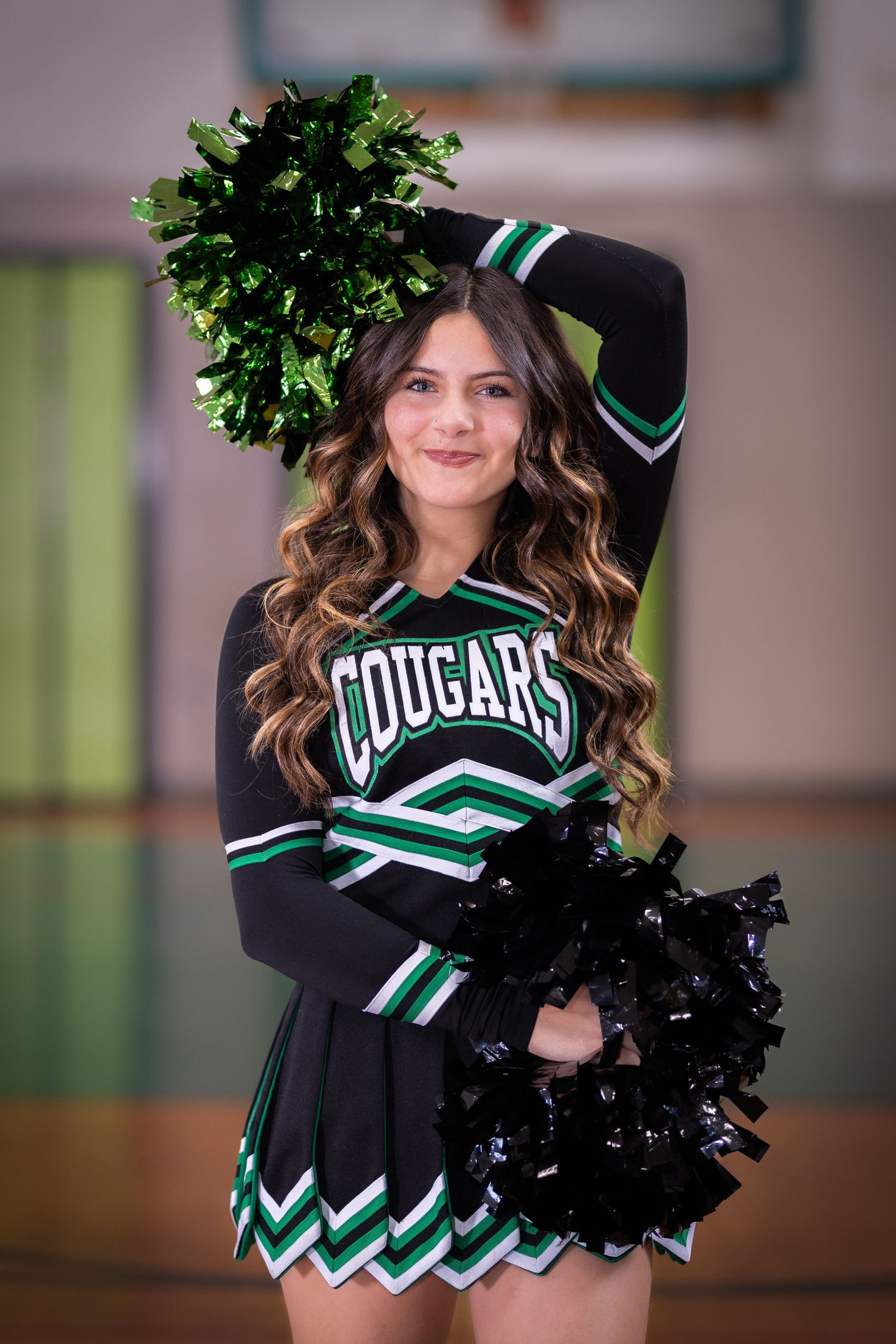 A smiling cheerleader in a Cougars uniform holds green and black pom-poms in a gym.
