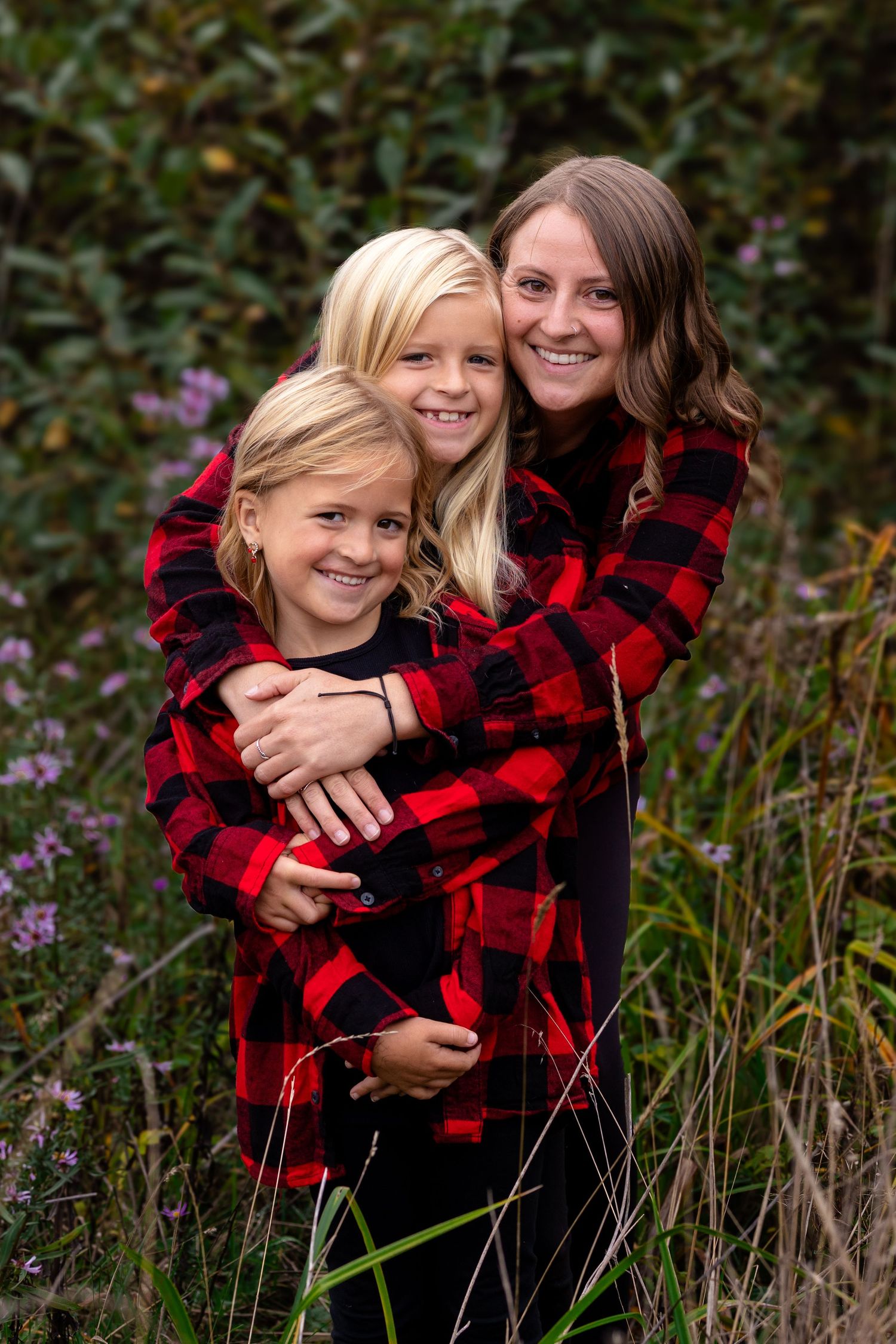 Three smiling girls in matching red plaid shirts embrace outdoors among green foliage and wildflowers.