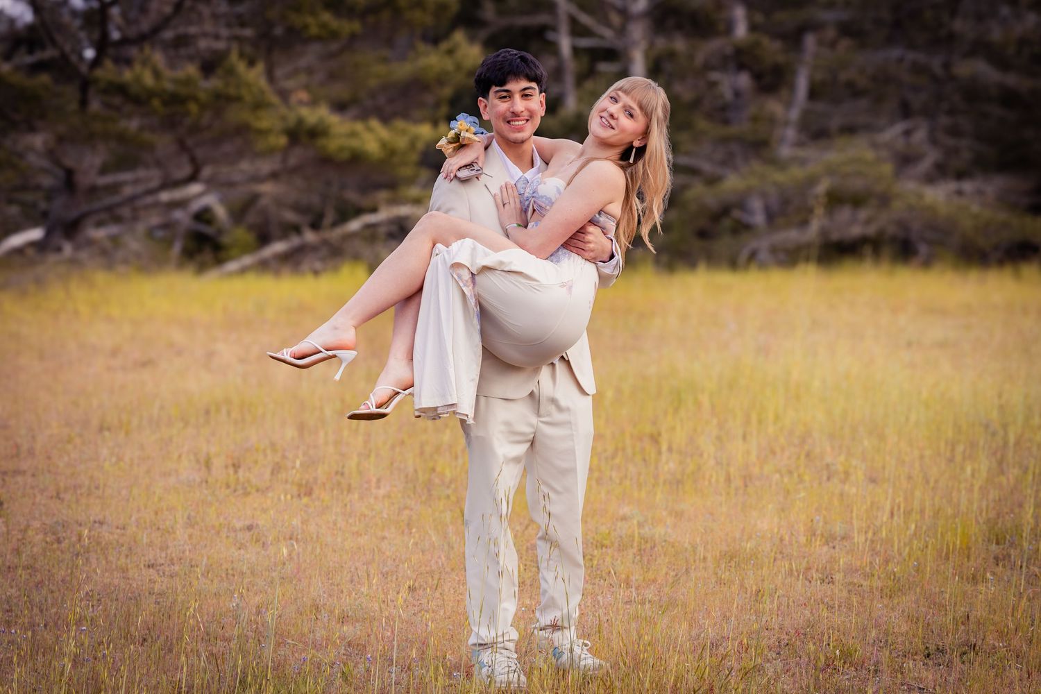 Couple posing in golden field, man carrying woman in his arms, surrounded by trees in natural outdoor setting.