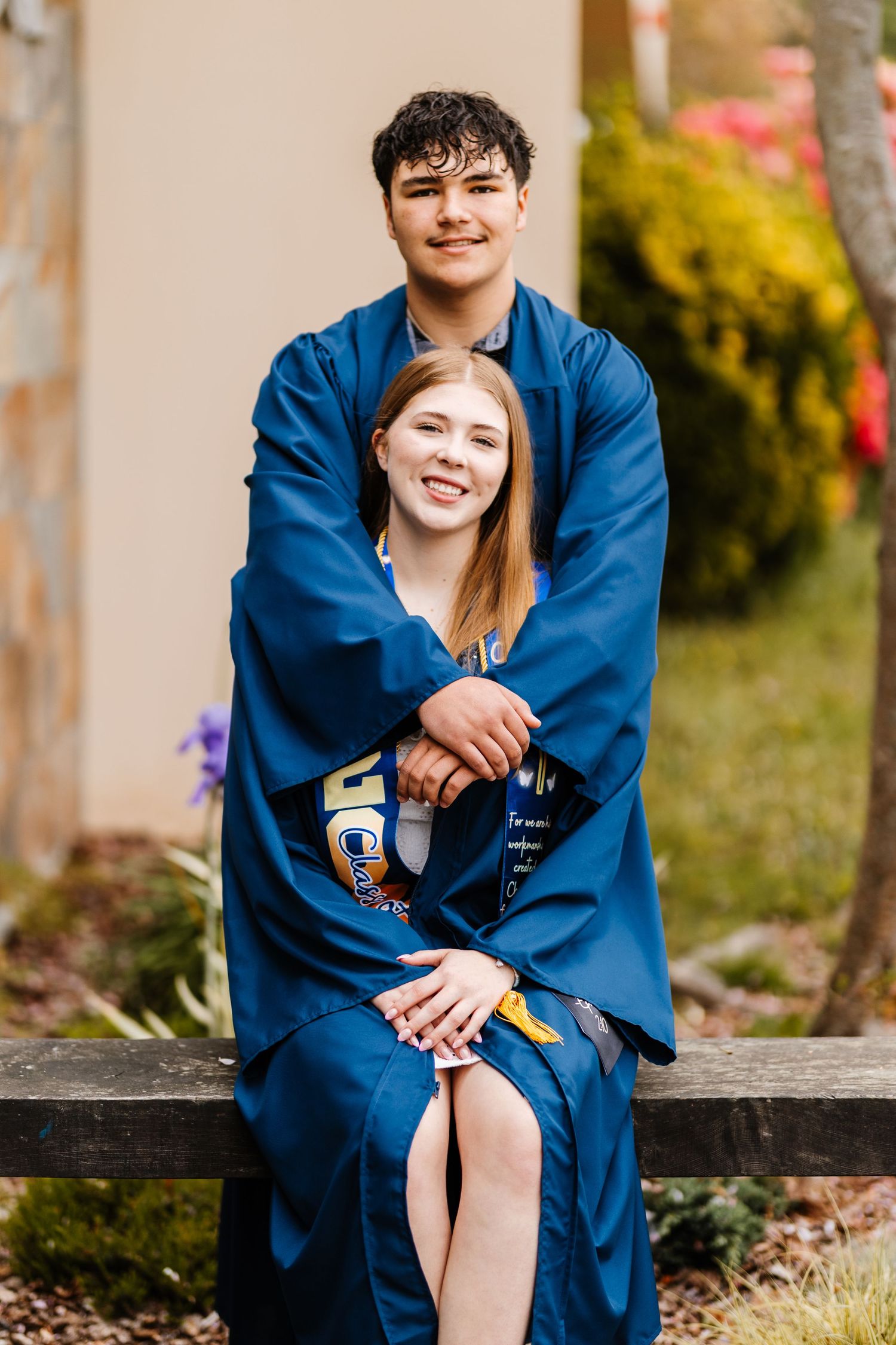 Two smiling graduates in blue robes pose together on a bench outdoors, surrounded by colorful spring flowers.