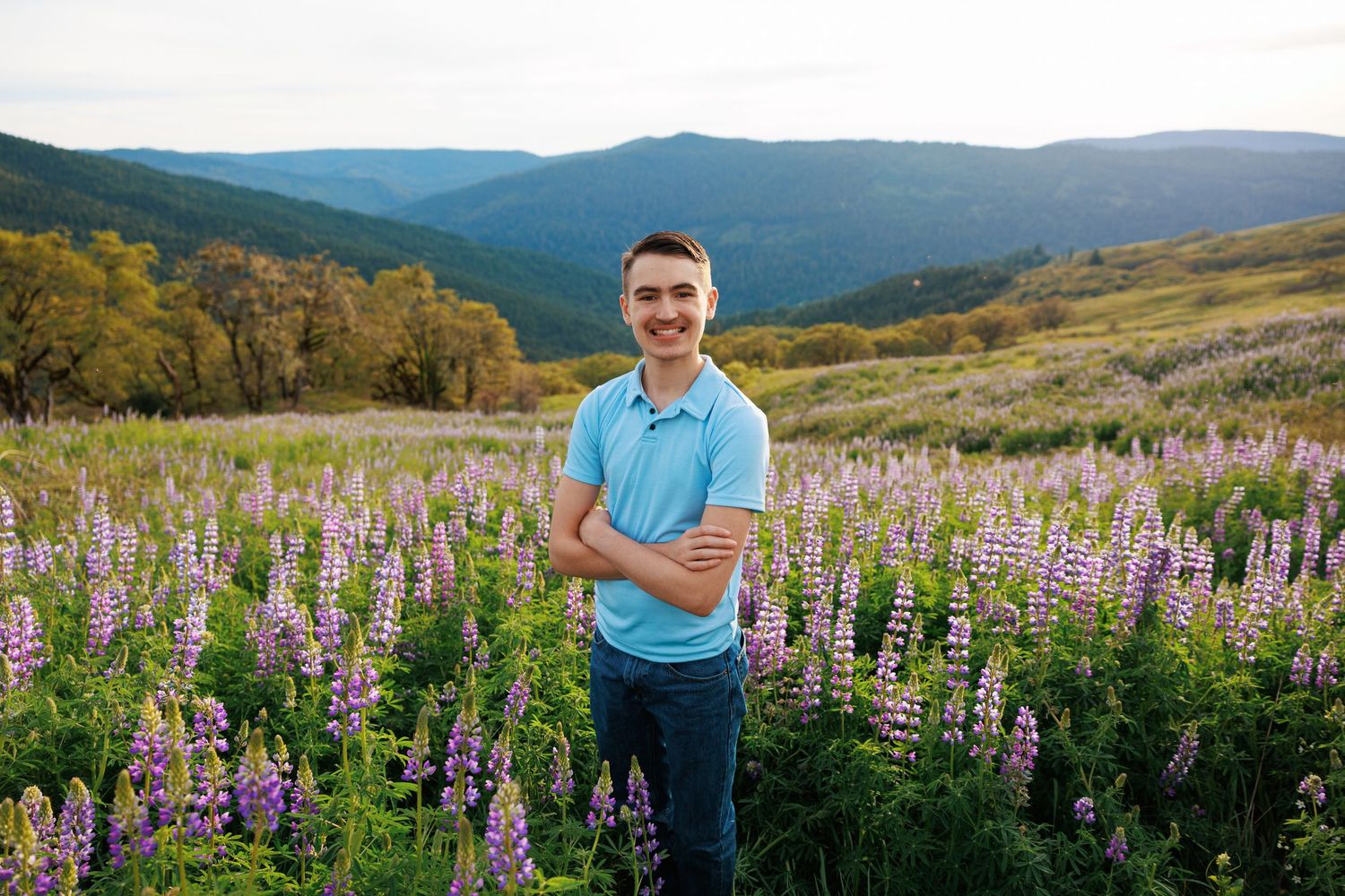 Young man in blue polo standing with crossed arms in purple lupine mountain field.