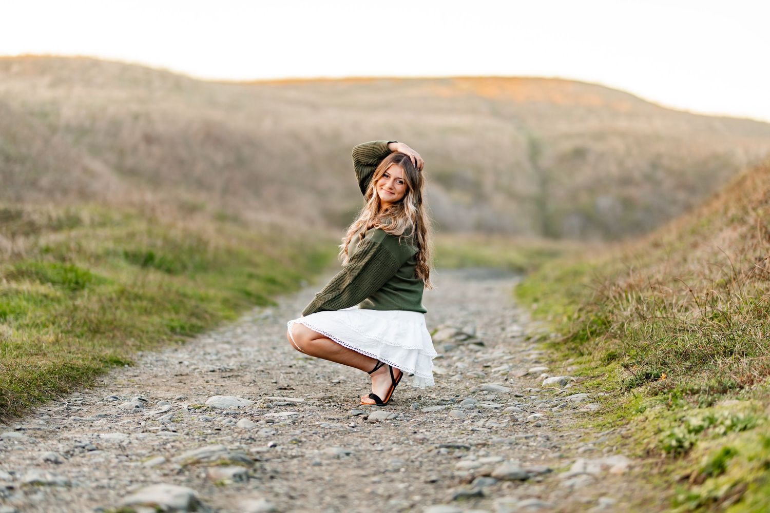 Blonde girl in green jacket and white skirt poses playfully on a gravel path in rolling hills.