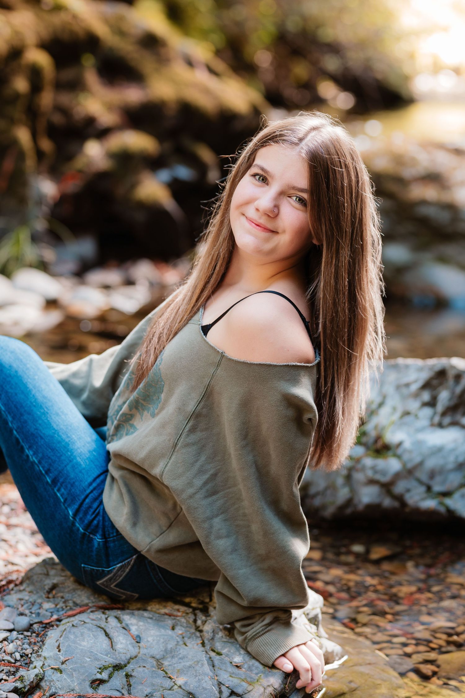 Teen girl with long brown hair sitting on rocks by a stream in olive off-shoulder sweater and jeans.