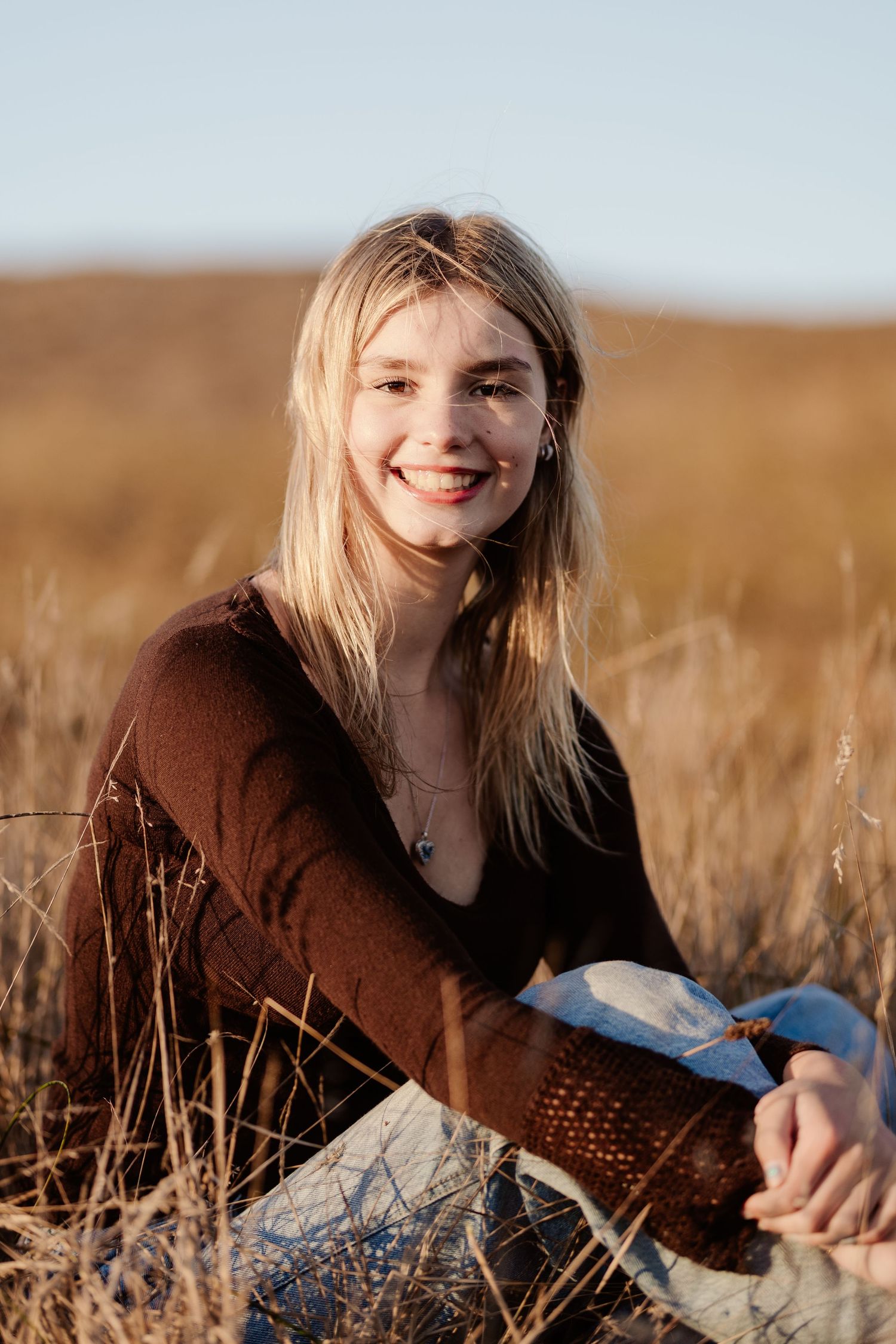 Smiling blonde teen girl sitting in golden dry grass field wearing brown top and jeans at sunset.