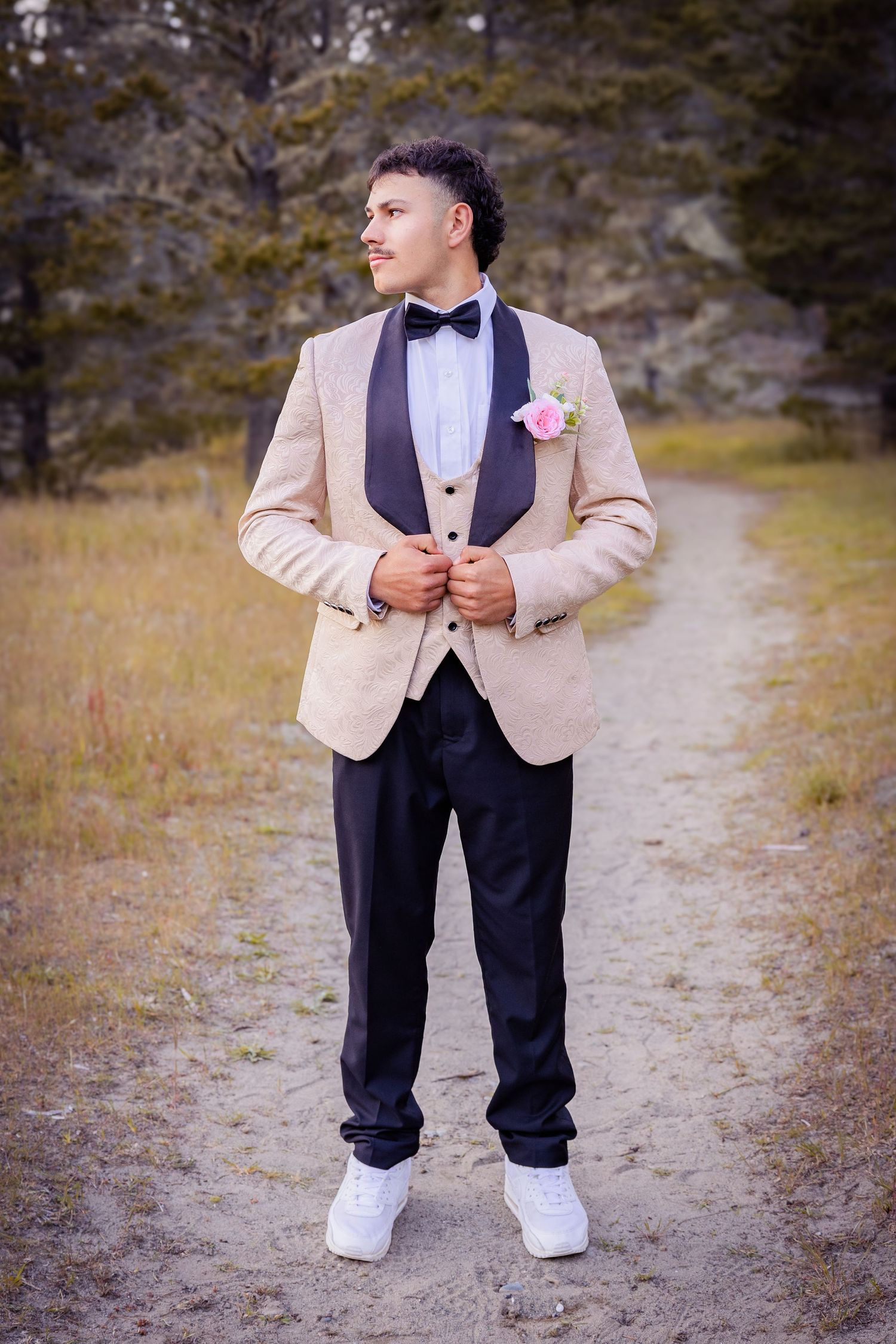 Young man in beige tuxedo jacket with black lapels, bow tie, and pink boutonniere poses on a dirt path outdoors.
