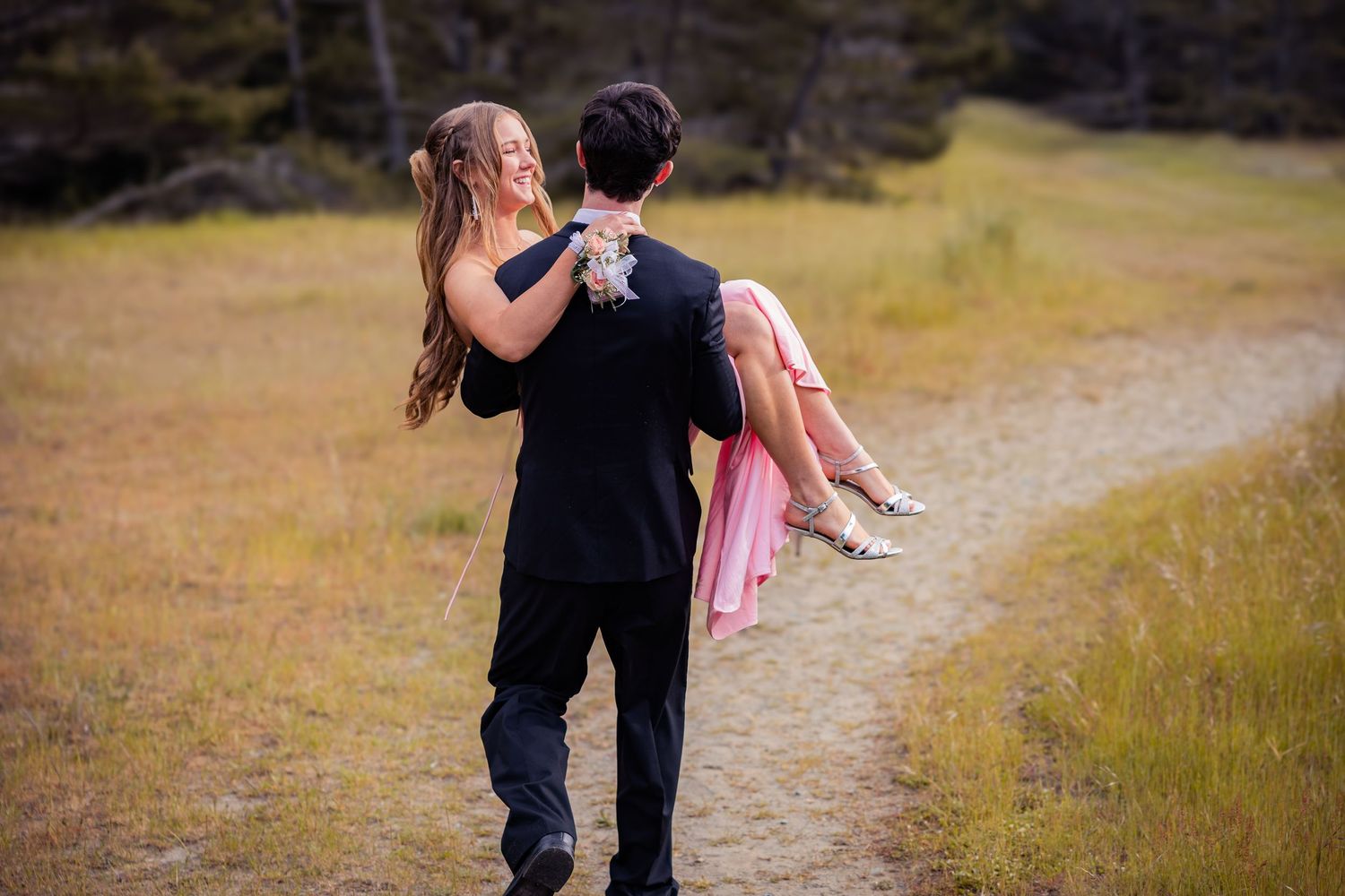 A young man in a black suit carries a woman in a pink dress down a dirt path through a golden meadow.