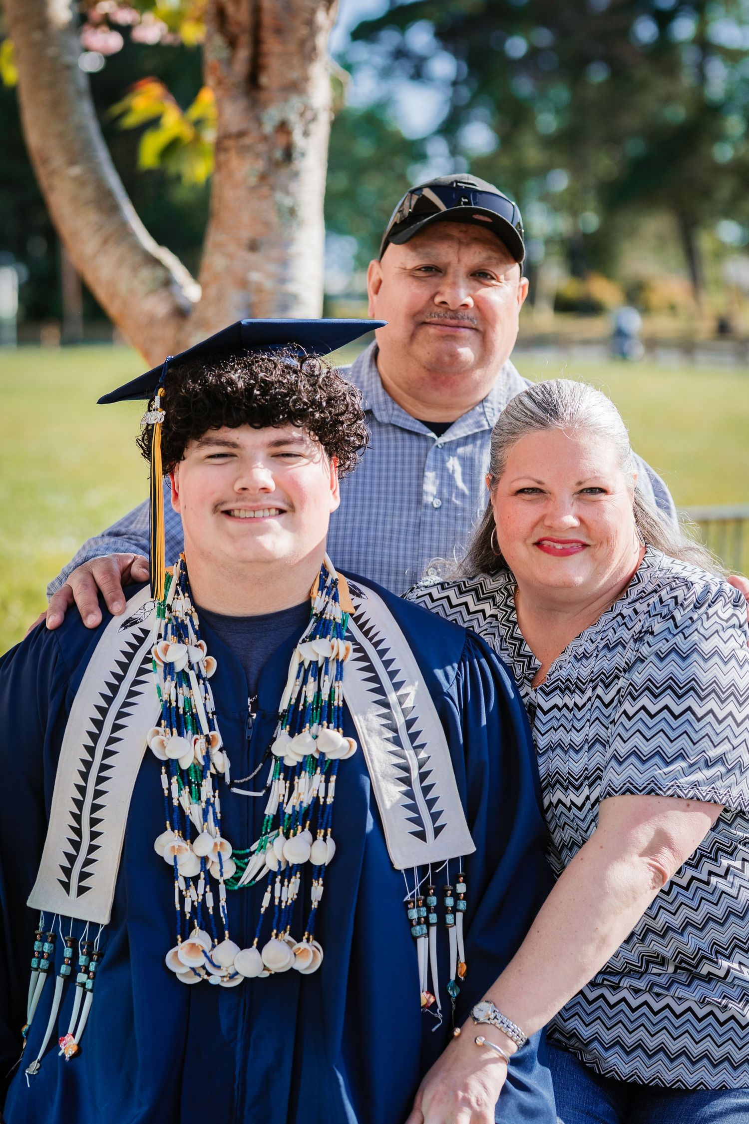 A smiling graduate in blue cap and gown poses with proud parents outdoors on a sunny graduation day.