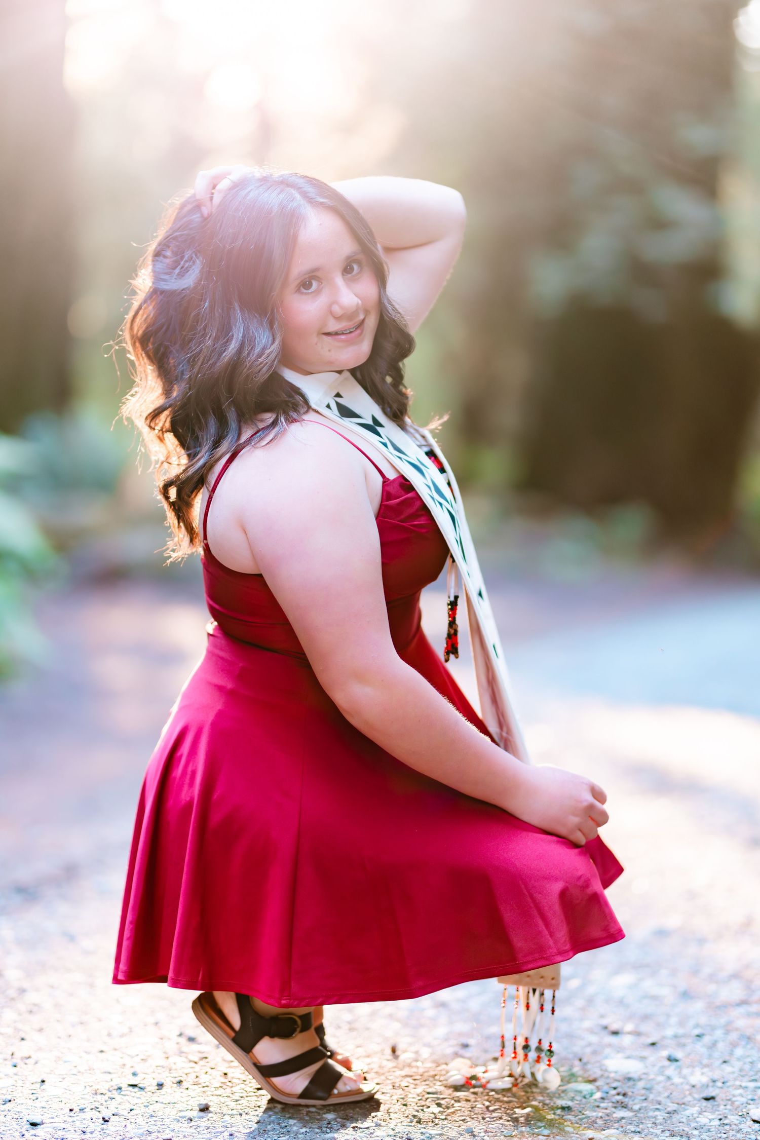 Young woman in red dress poses gracefully on sunlit forest path, smiling over shoulder with hand in hair.
