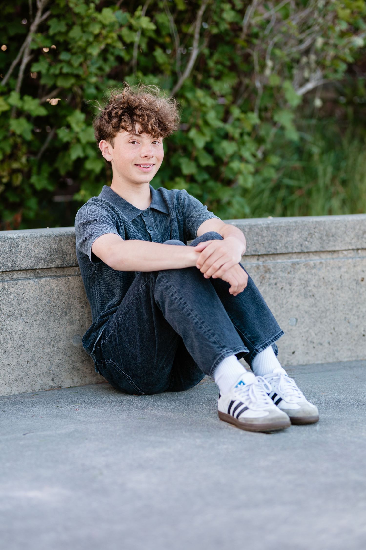 Smiling teen boy with curly hair sits against stone wall outdoors, wearing gray polo, jeans, and white Adidas sneakers.