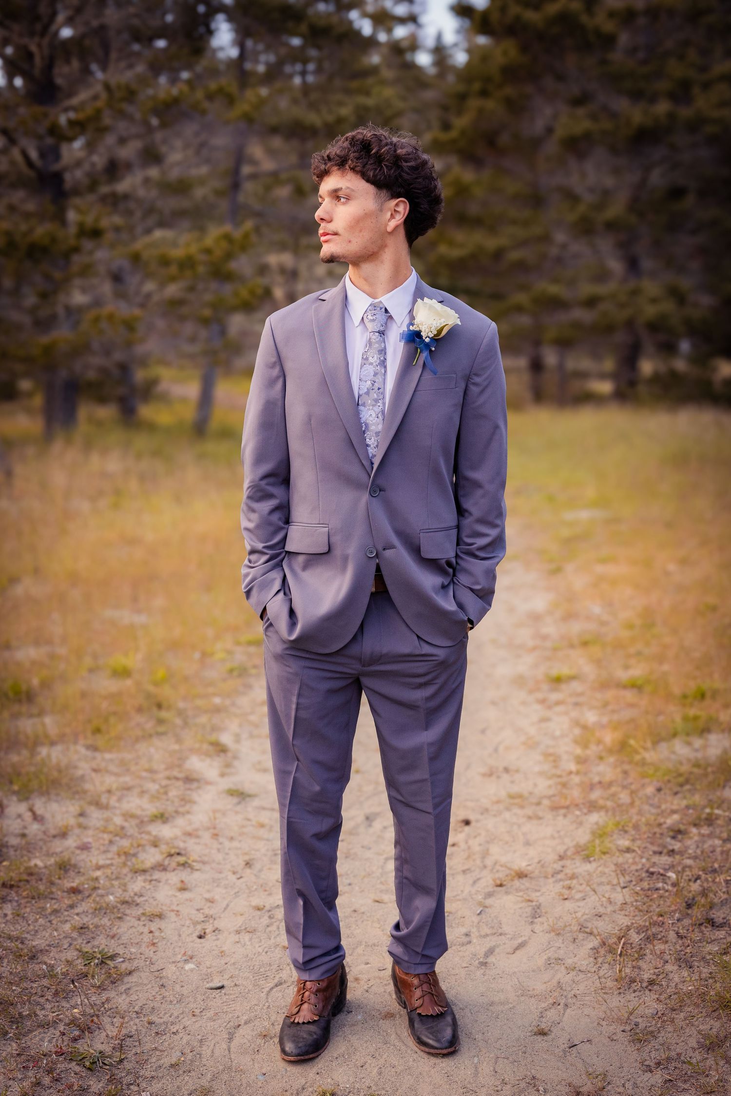 A groom in a gray suit with floral tie and boutonniere stands on a dirt path surrounded by trees.