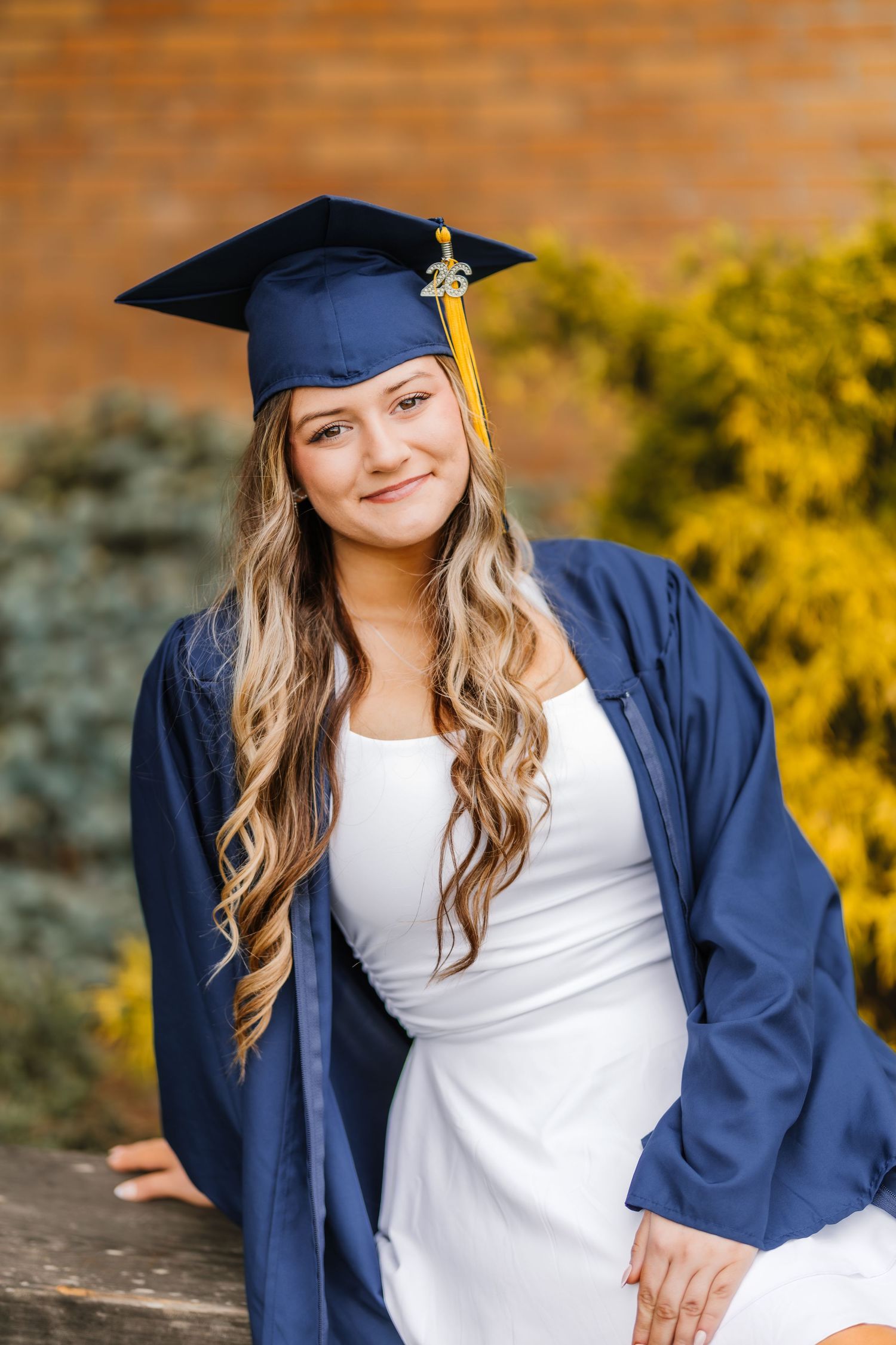Young woman in navy graduation cap and gown smiling outdoors near brick wall.