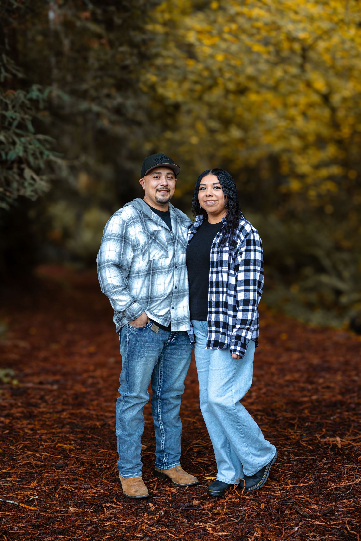 Smiling couple in coordinating plaid flannel shirts pose together on a scenic autumn redwood forest pathway.