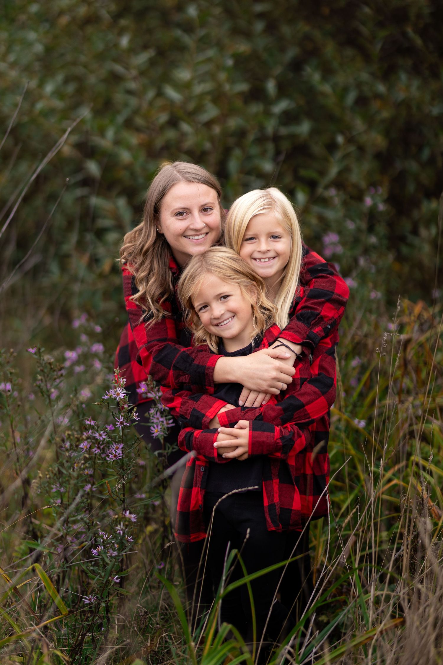 Mother hugging two blonde daughters wearing matching red plaid shirts in autumn field.