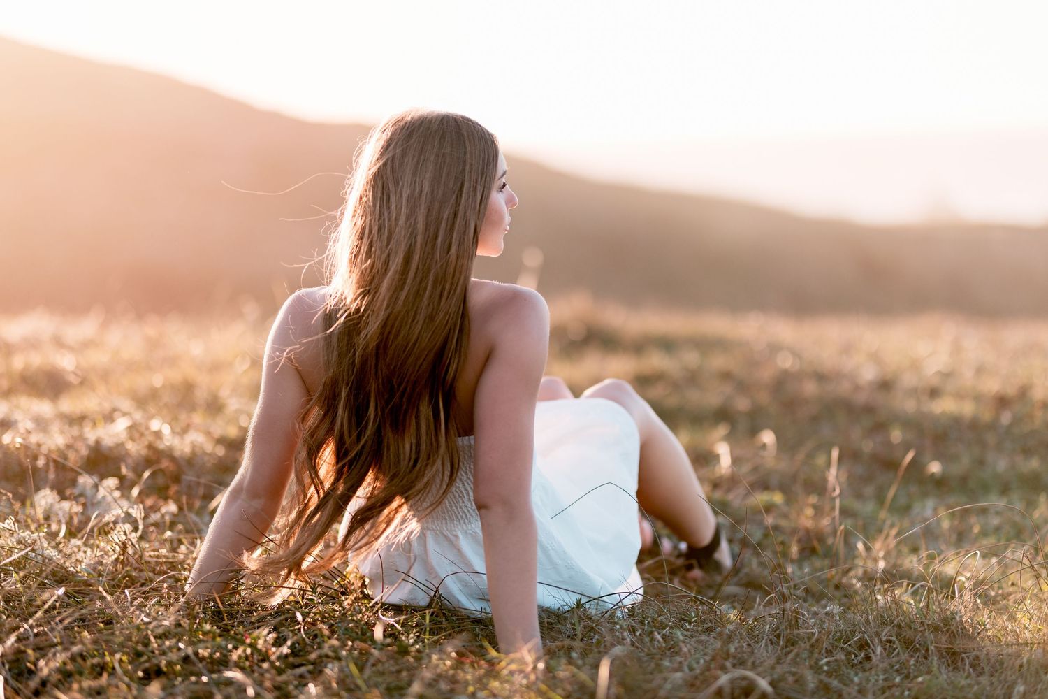 Young woman in white dress sits in golden meadow at sunset, gazing at scenic mountain landscape.