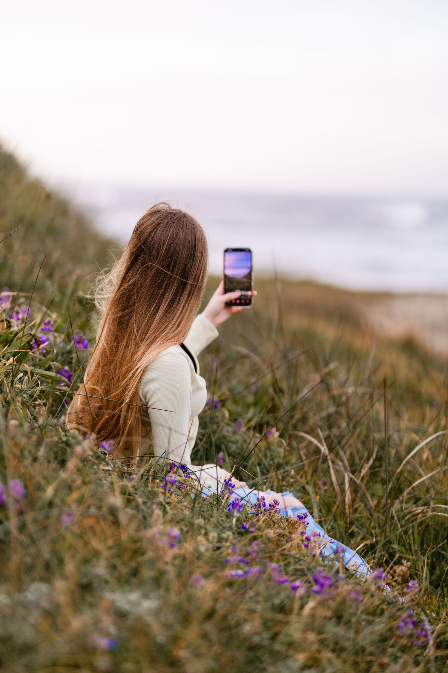 Young woman with long hair photographing coastal scenery with smartphone in a flower field.