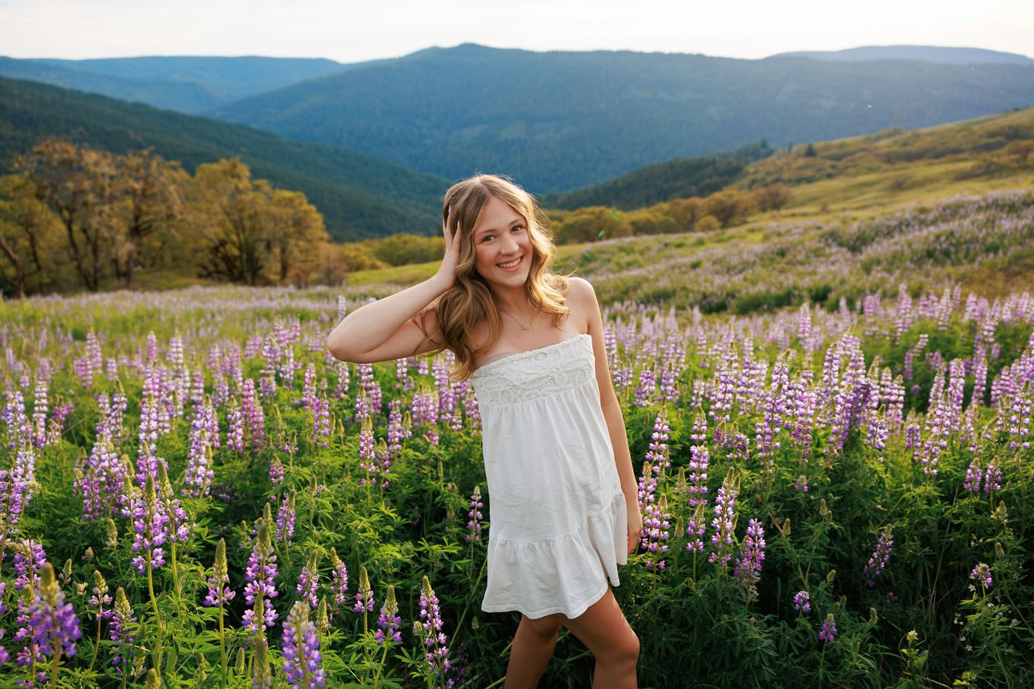 Teenage girl in white dress smiling in purple lupine field with mountain backdrop.