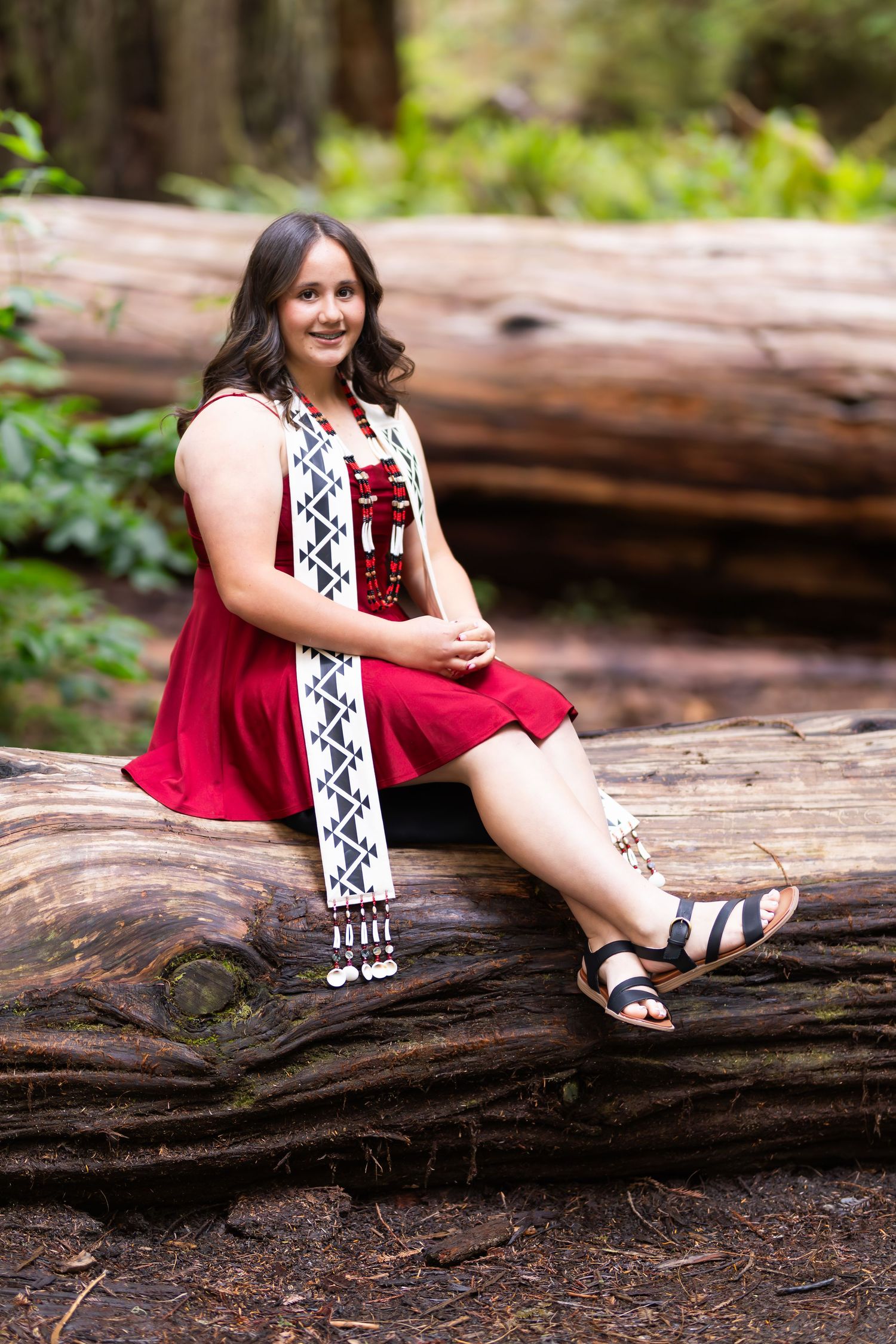 Young woman in red dress and beaded sash sits on fallen log in lush forest setting.