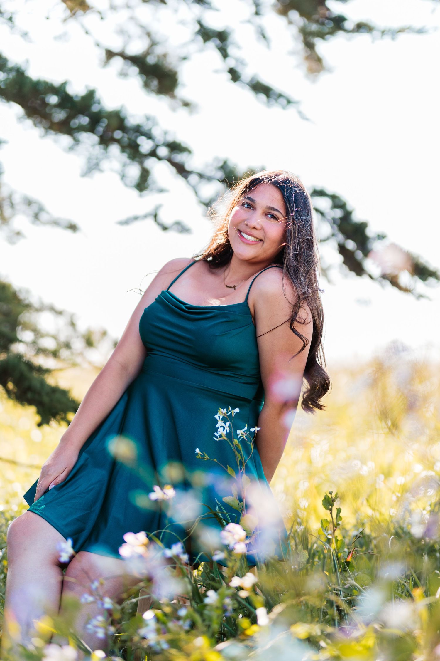Happy young woman in teal dress posing in a sunny wildflower meadow under a tree.